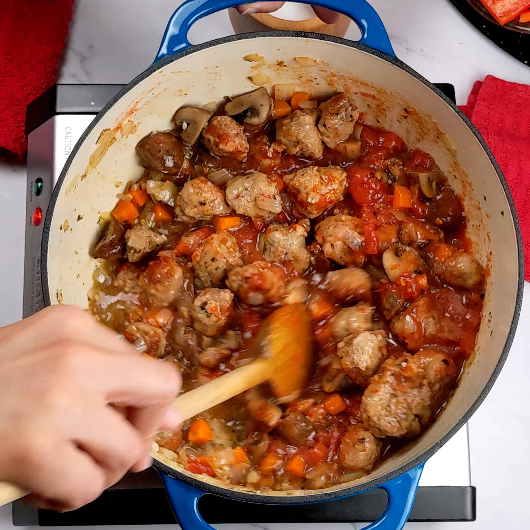 browned italian sausage with tender mushrooms, carrots, onion and garlic with crushed tomatoes and harrissa paste combined in a dutch oven being mixed with a wooden spoon