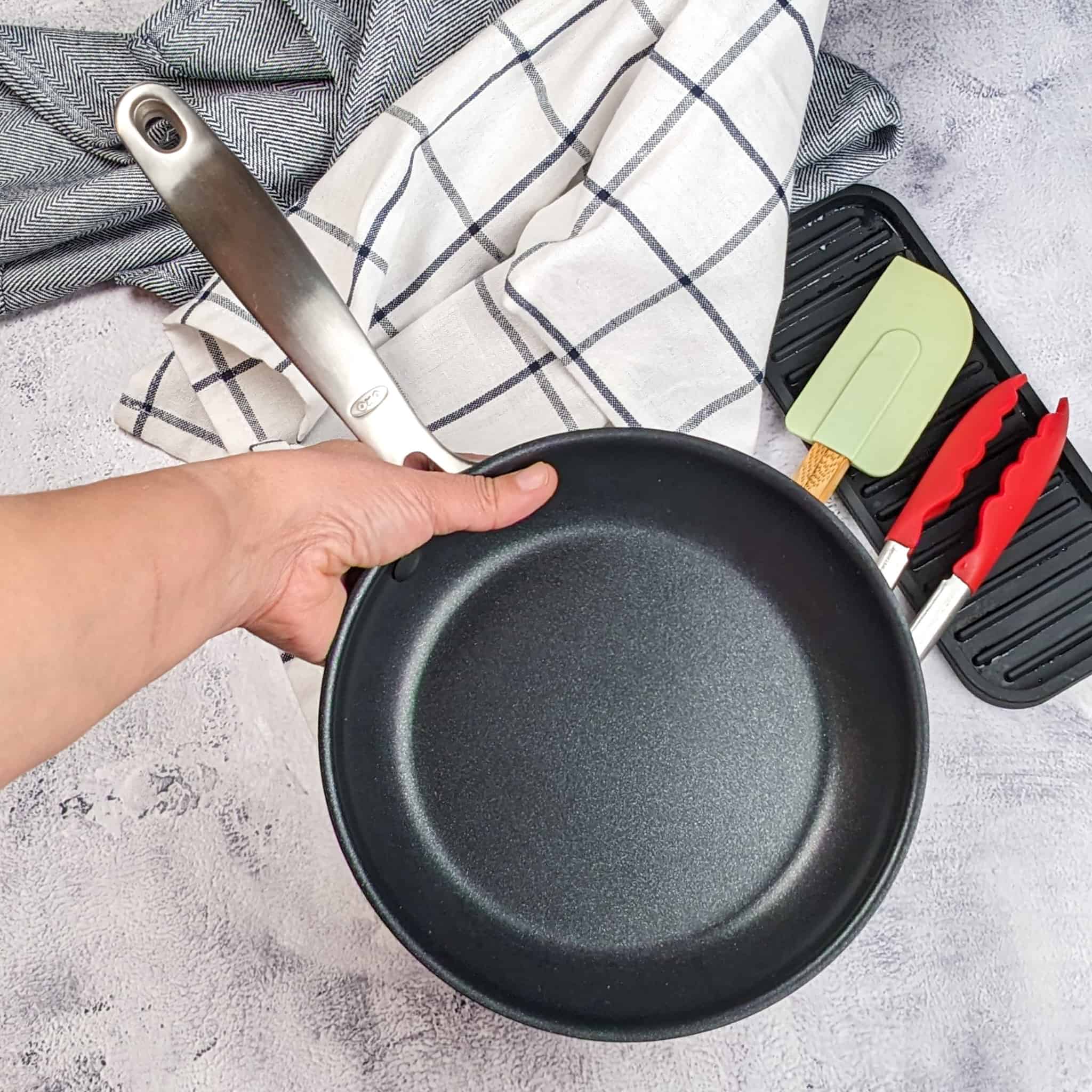 The OXO Good Grips Pro 8" frying pan skillet held in the hand over a background of towels, silicone resting tray, a KitchenAid silicone spatula and a pair of KitchenAid silicone tongs.