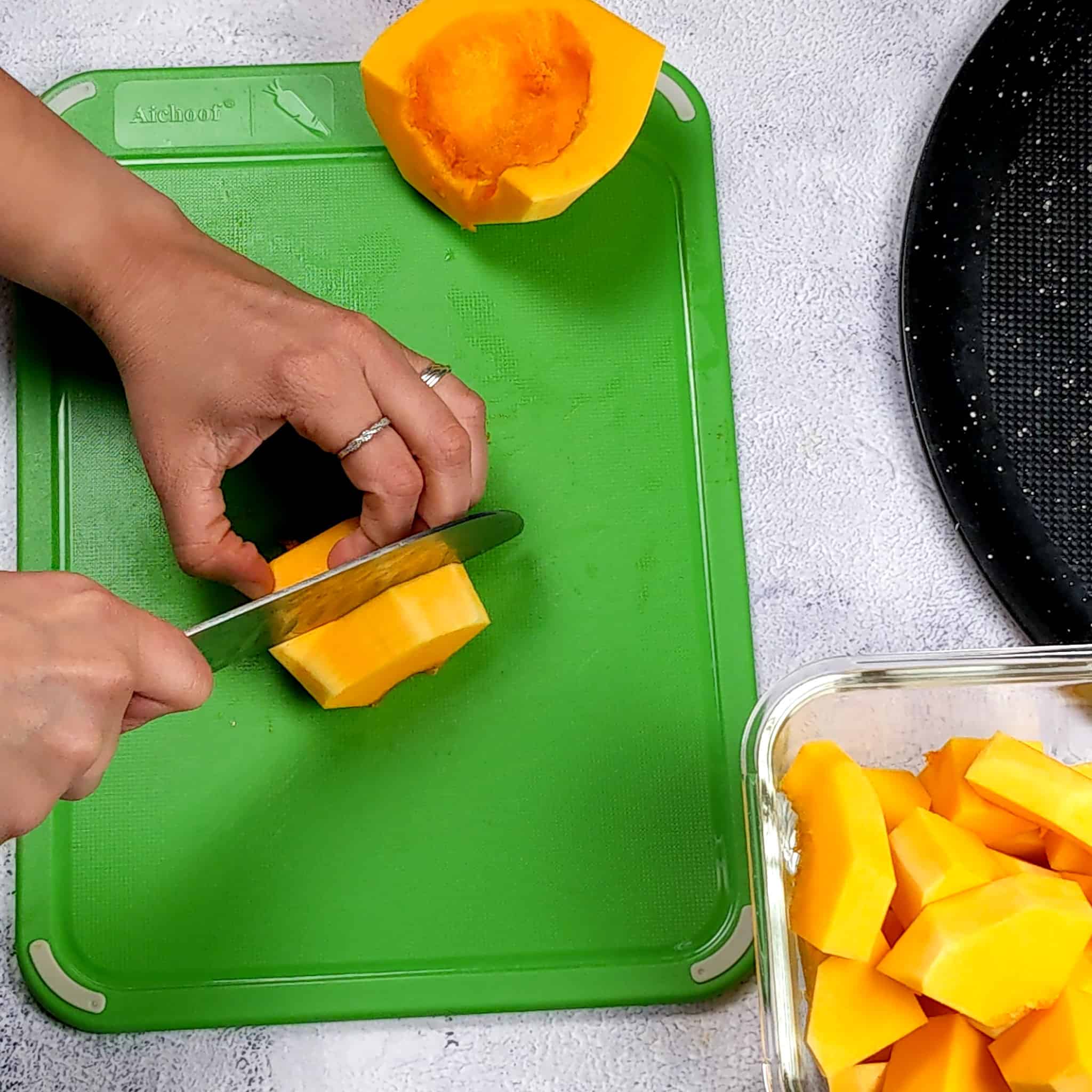 a peeled butternut squash being cut into thick slices on a small color coded cutting board with a knife next to a container of already sliced pieces.