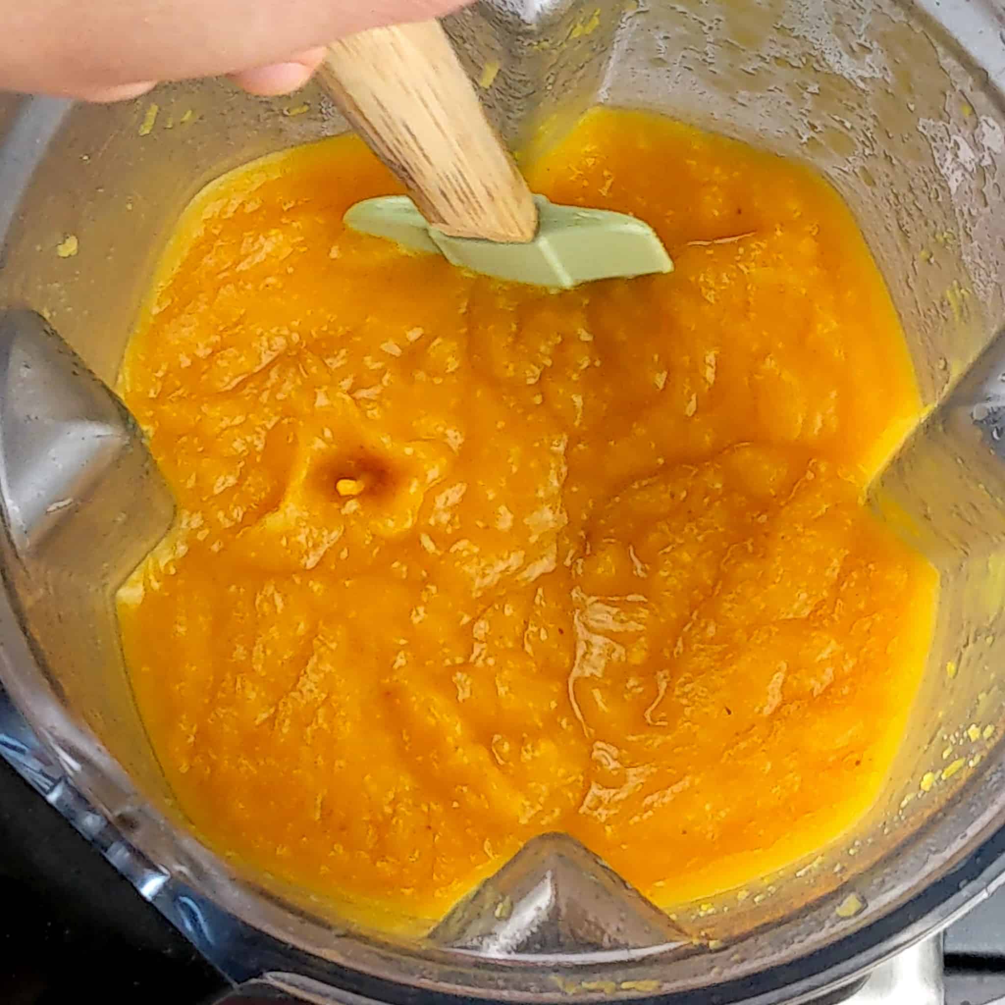 pureed roasted butternut squash in a blender container being scrapped and mixed with a silicone spatula with a wooden handle