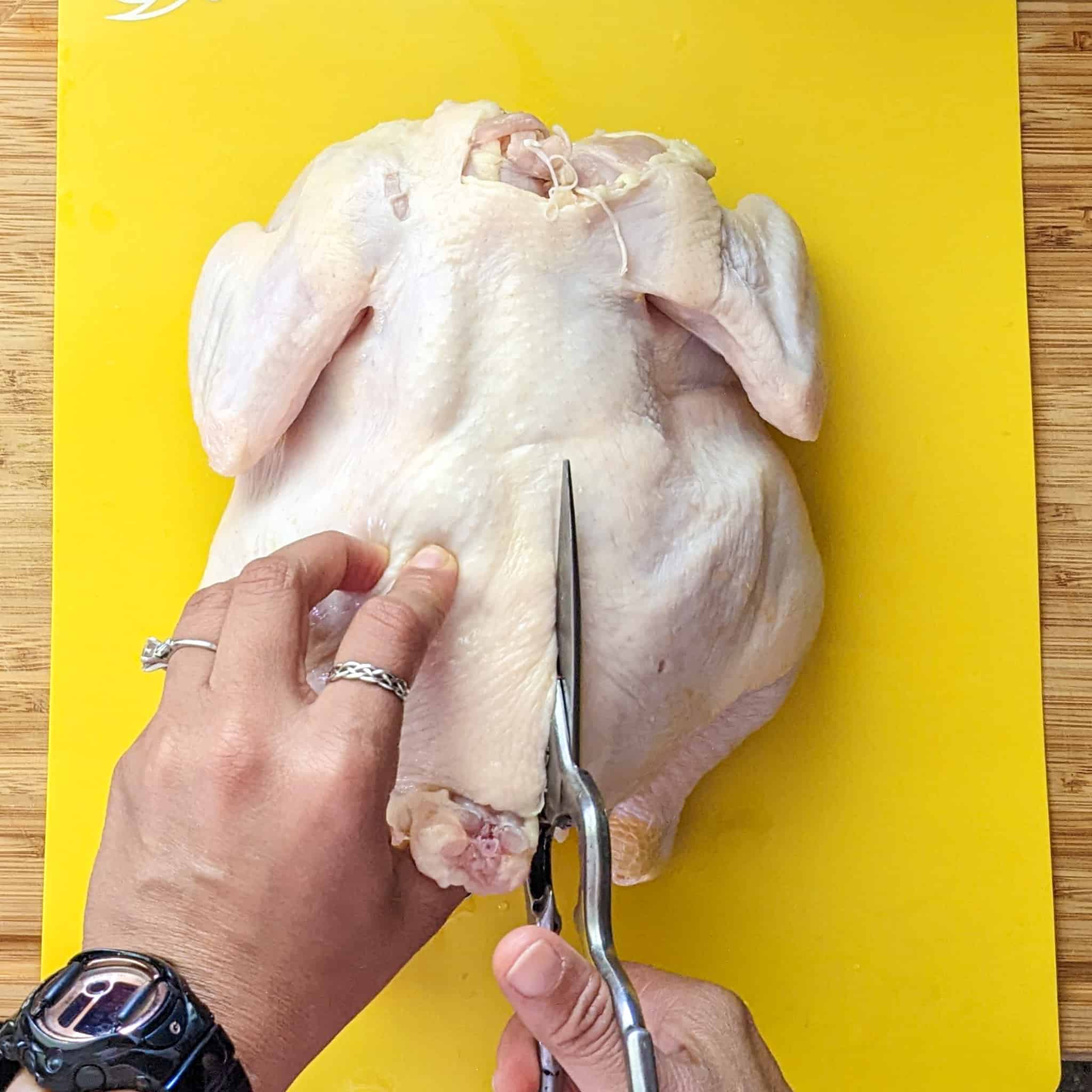 whole chicken on the cutting board being cut with kitchen shears from the bottom up on the right side of the chicken's backbone.