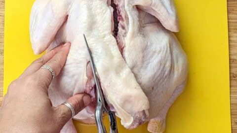 whole chicken on the cutting board being cut with kitchen shears from the bottom up on the left side of the chicken's backbone.
