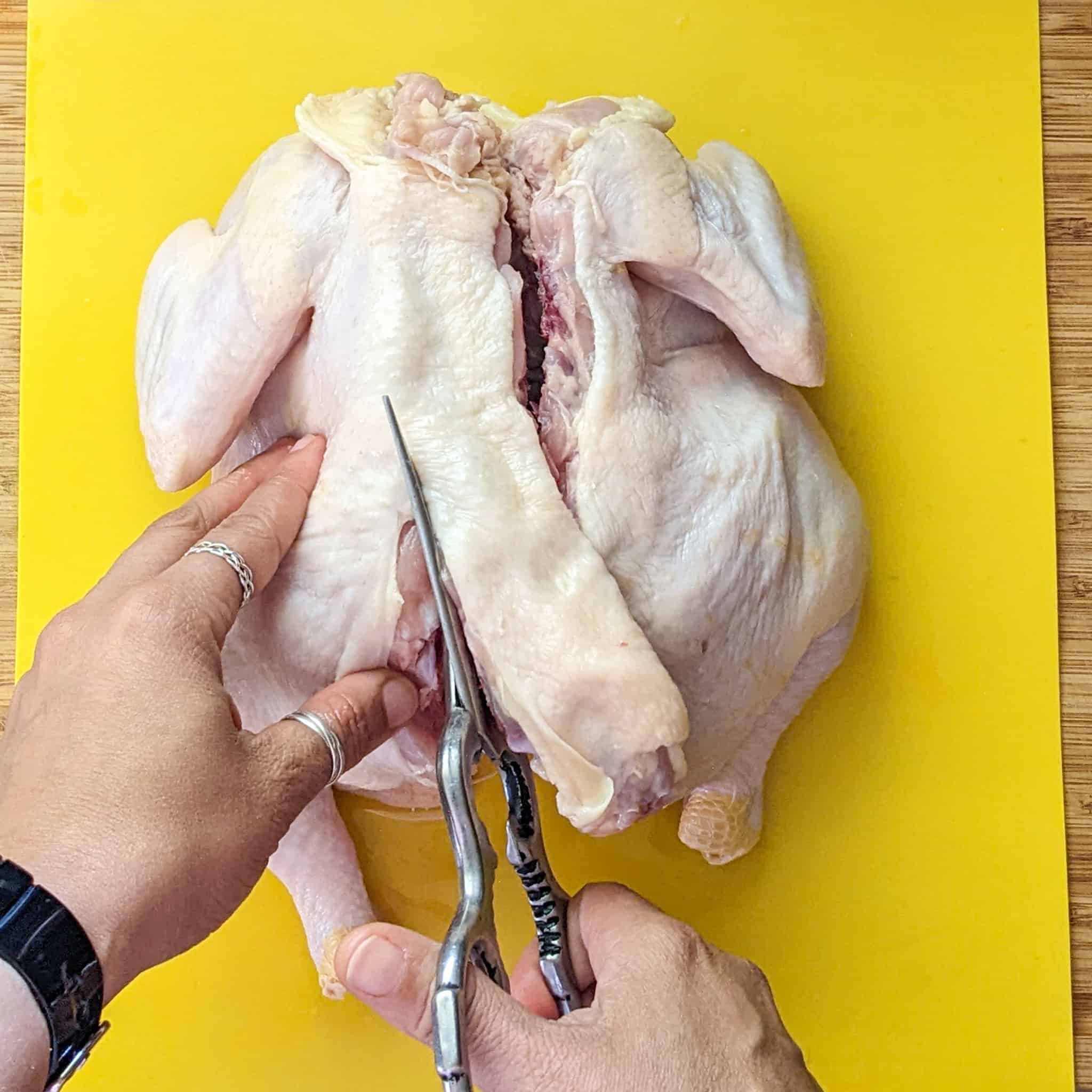 whole chicken on the cutting board being cut with kitchen shears from the bottom up on the left side of the chicken's backbone.
