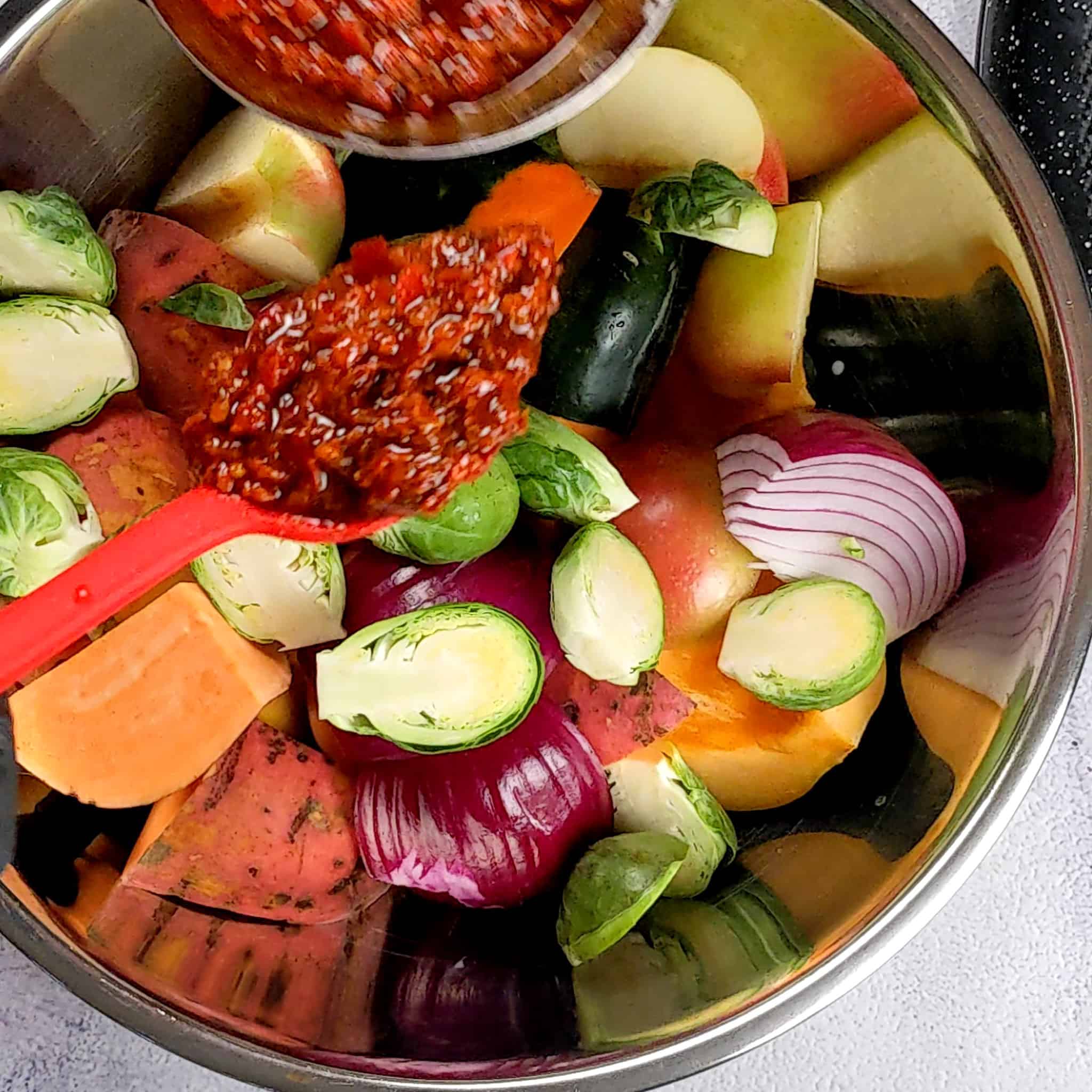 acorn squash, sweet potatoes, red onions, brussel sprouts, and apples cut into large chunks in a large metal mixing bowl with a GIR silicone table spoon being held right above the bowl with wet rub