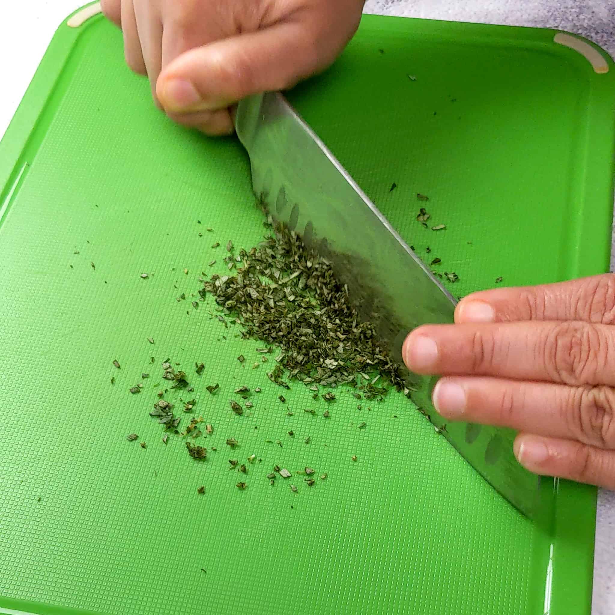hands chopping fresh rosemary on a plastic color coded cutting board with a knife