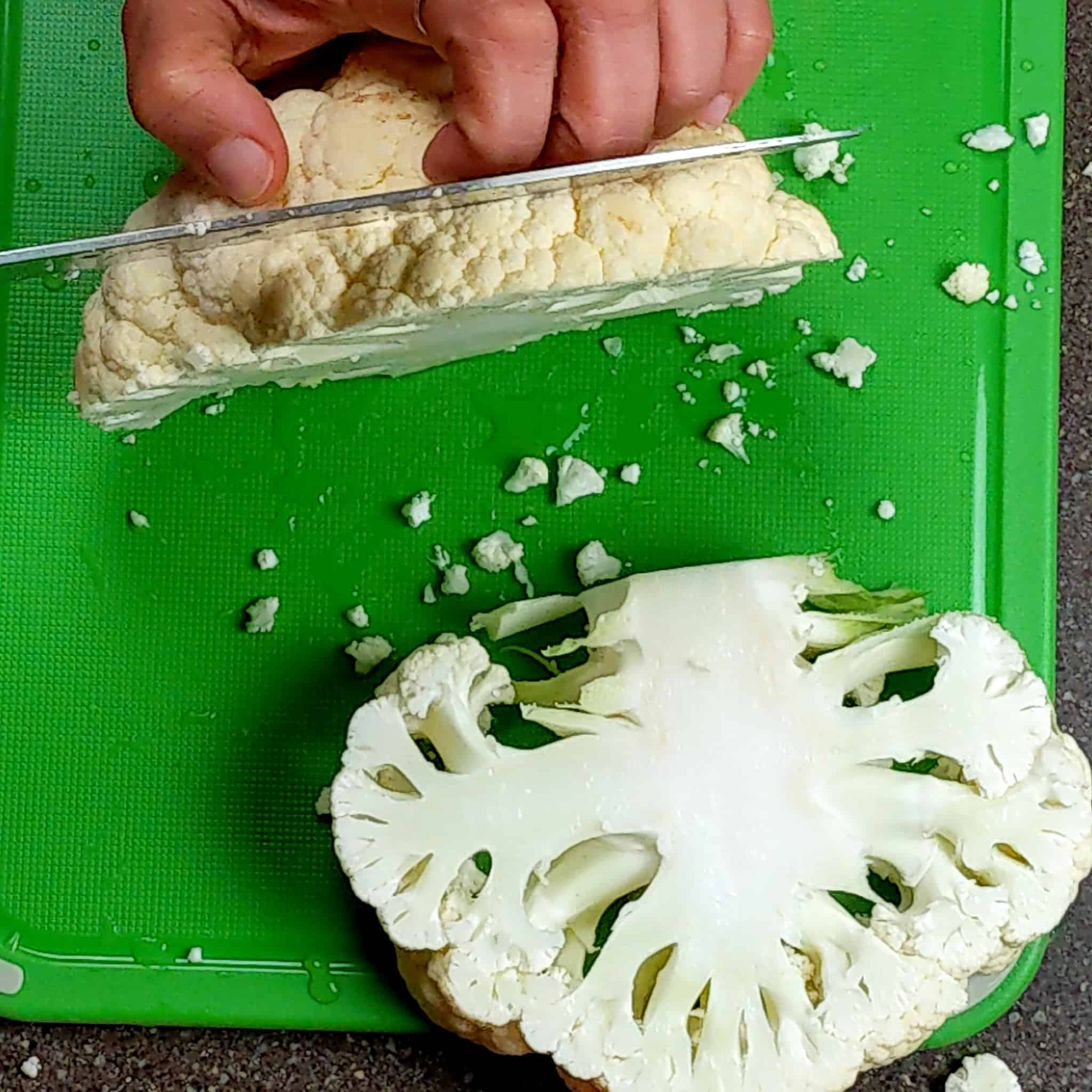 knife cutting the already cut cauliflower head with one raw cauliflower steak lays on the cutting board