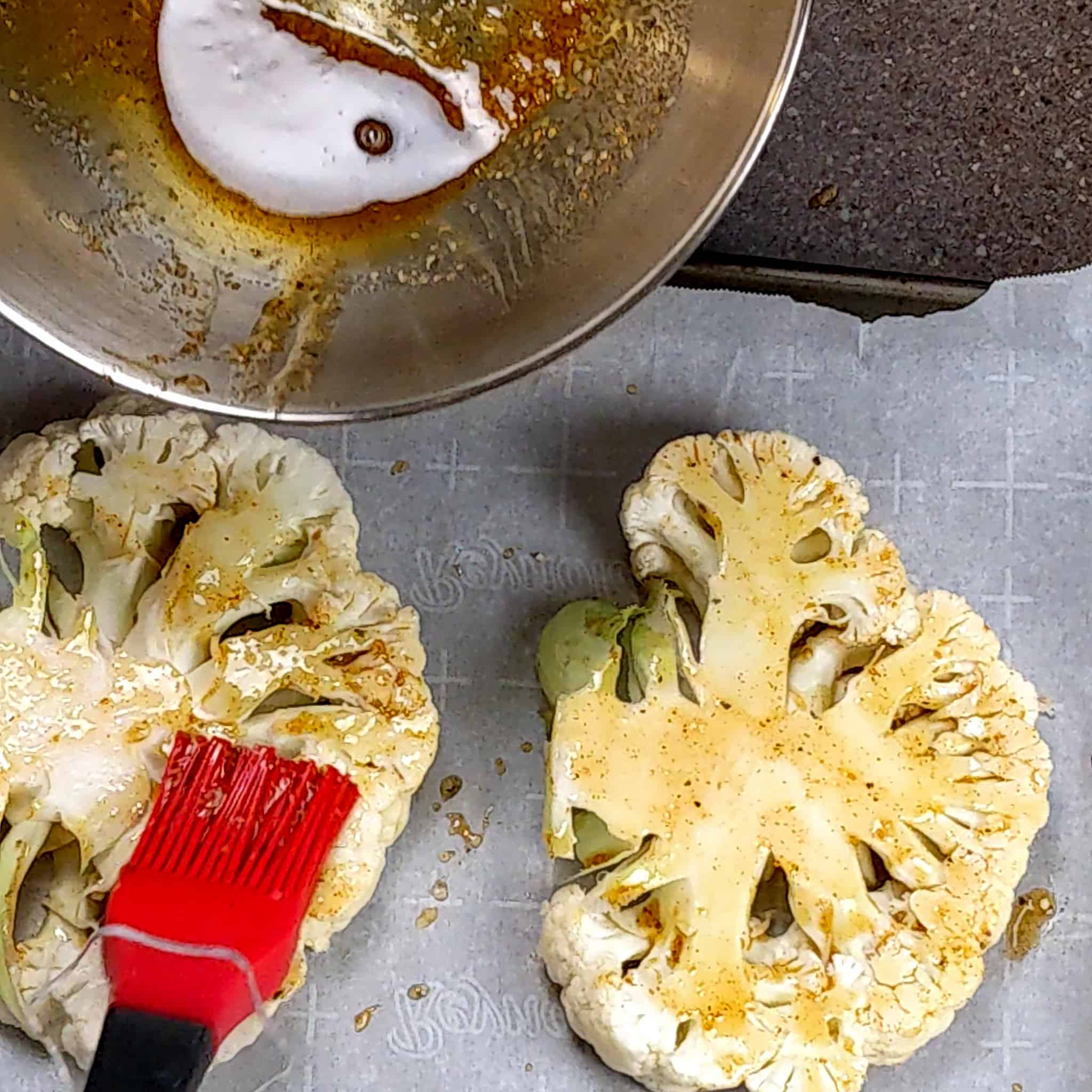 silicone pastry brush brushing the top of the cauliflower steaks on a sheet pan with parchment paper