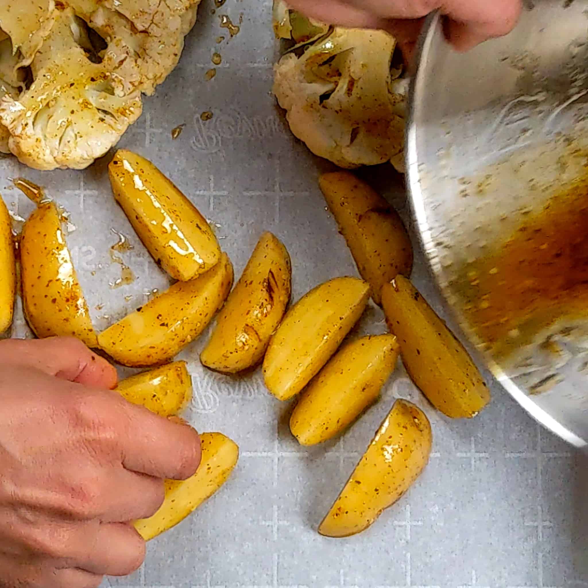 wet rub seasoning potato wedges being laid on a sheet pan with parchment paper next to the seasoned cauliflower steaks