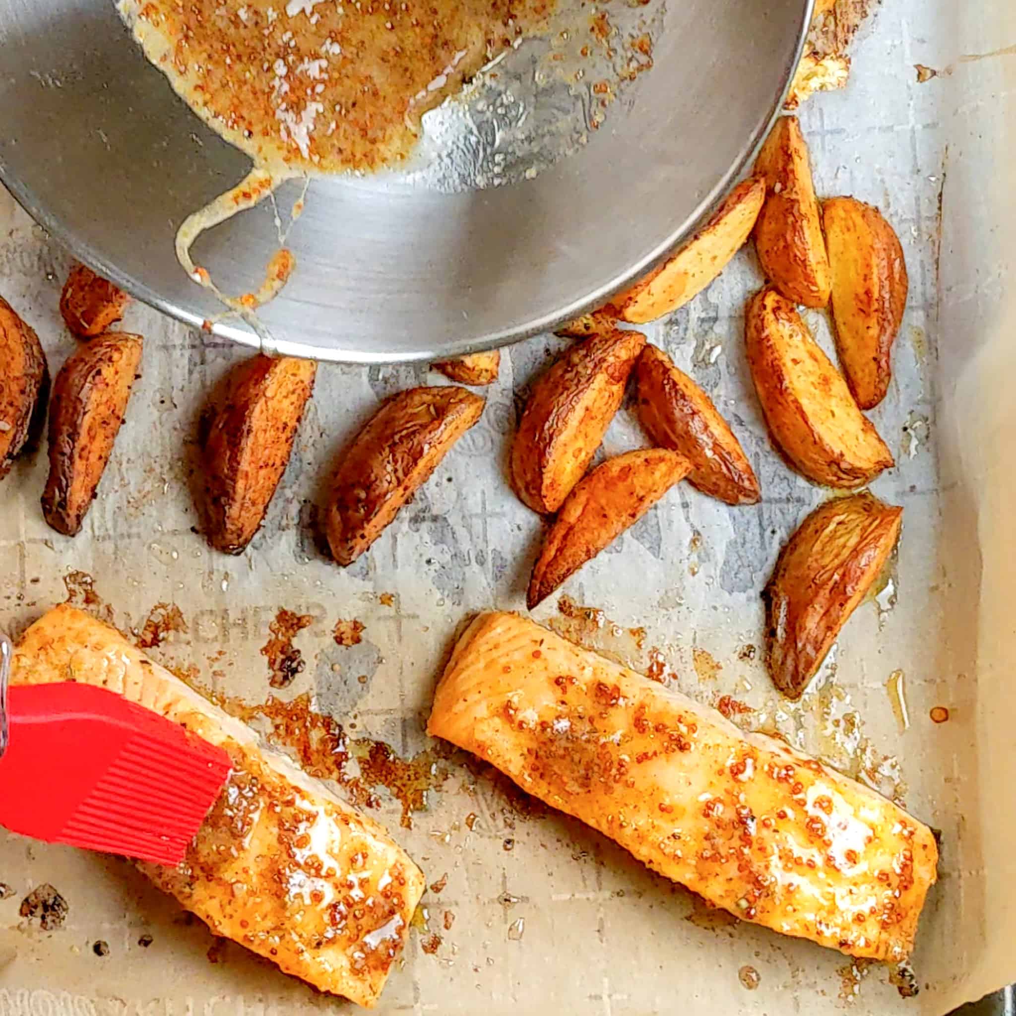 baked salmon being brushed with the honey dijon mustard on the sheet pan with the baked potato wedges