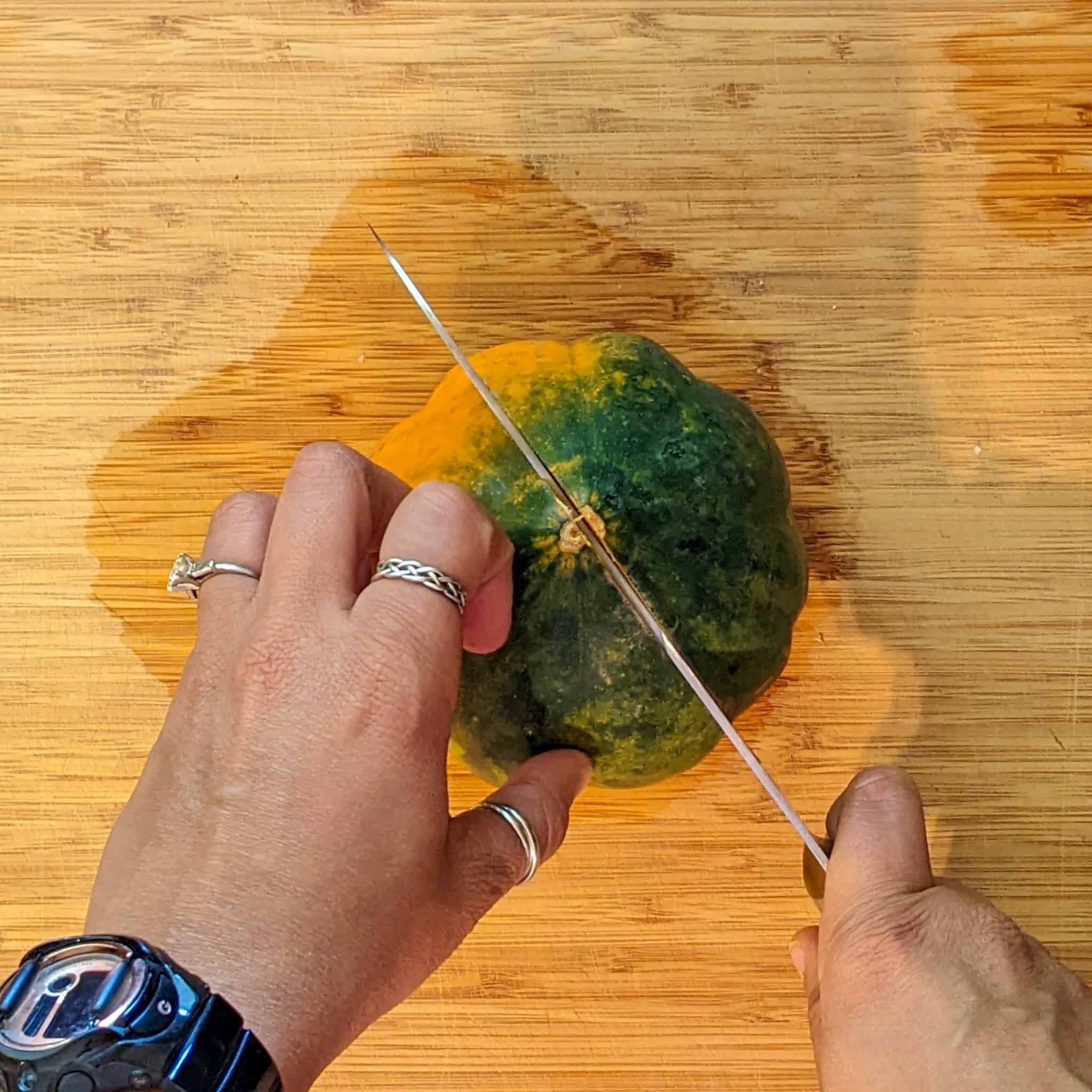 top view of an acorn squash being cut into have vertically with a knife on a cutting board