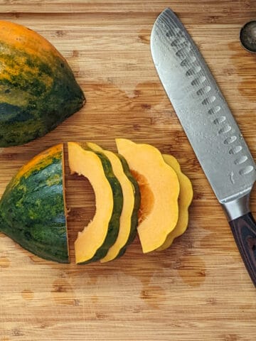 two halved acorn squash skin side up. one in whole while the other has been cut into sliced rings half way through laying on a cutting board next to a knife