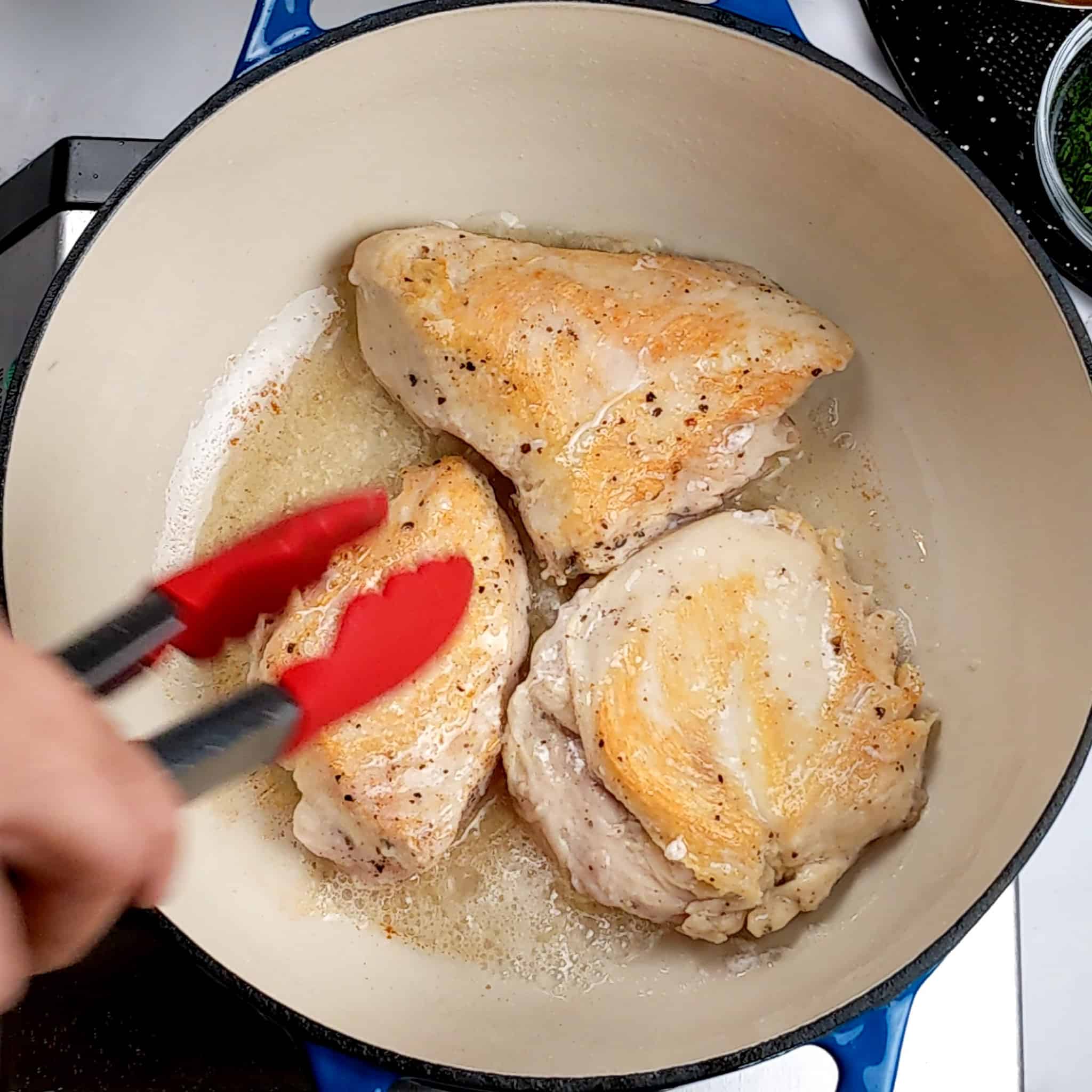 chicken breast searing in a Dutch oven in oil and being flipped with a pair of silicone tongs