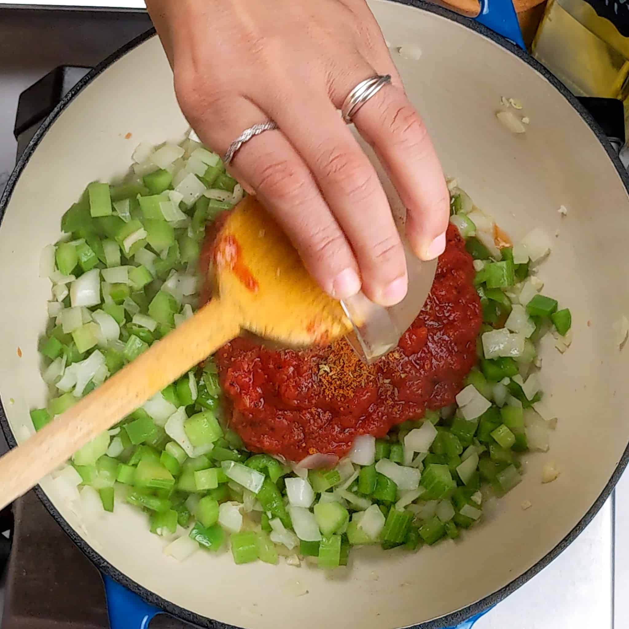 spices and crushed tomatoes being poured on top of sweated diced onion, bell pepper, celery and garlic.