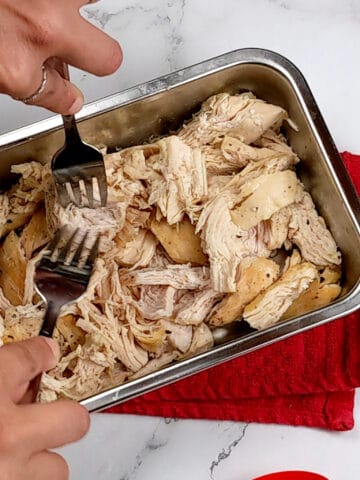 chicken being shredded with two forks in a metal rectangular container.