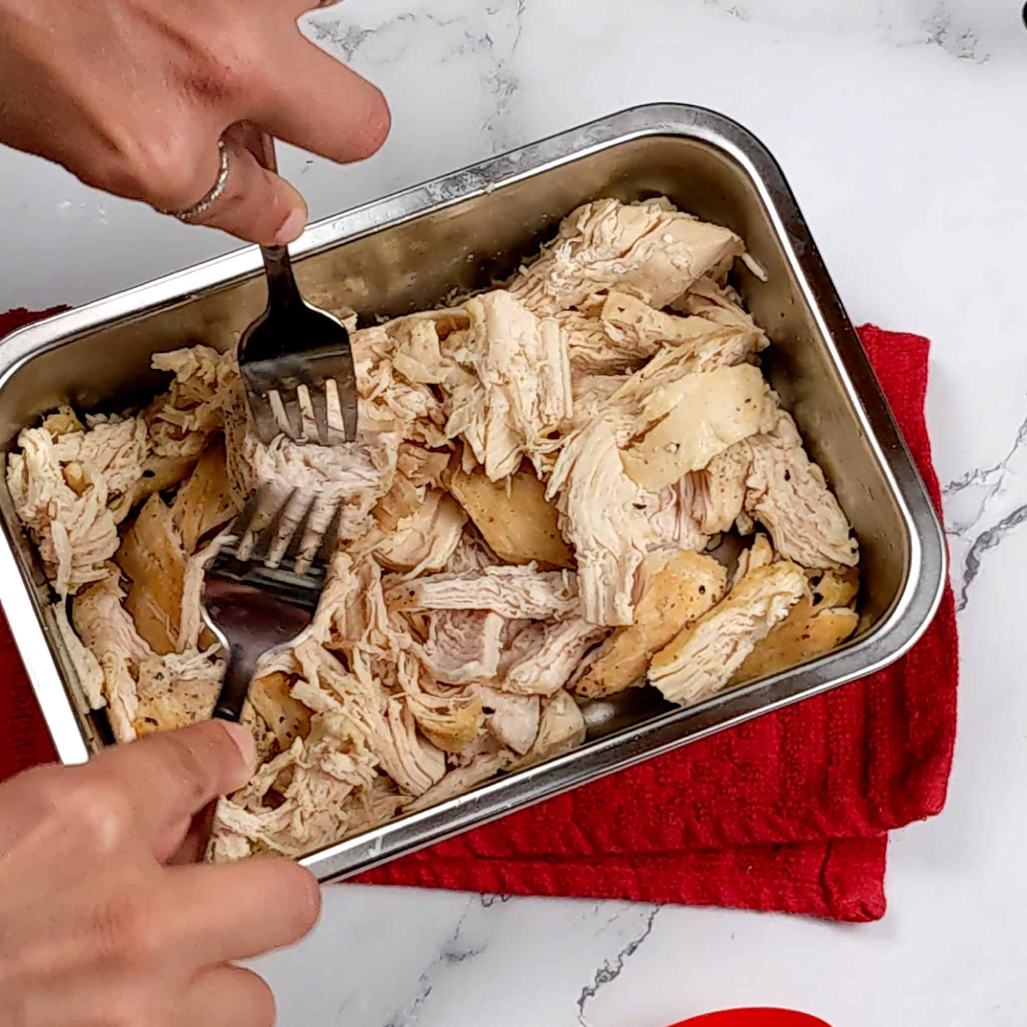 chicken being shredded with two forks in a metal rectangular container