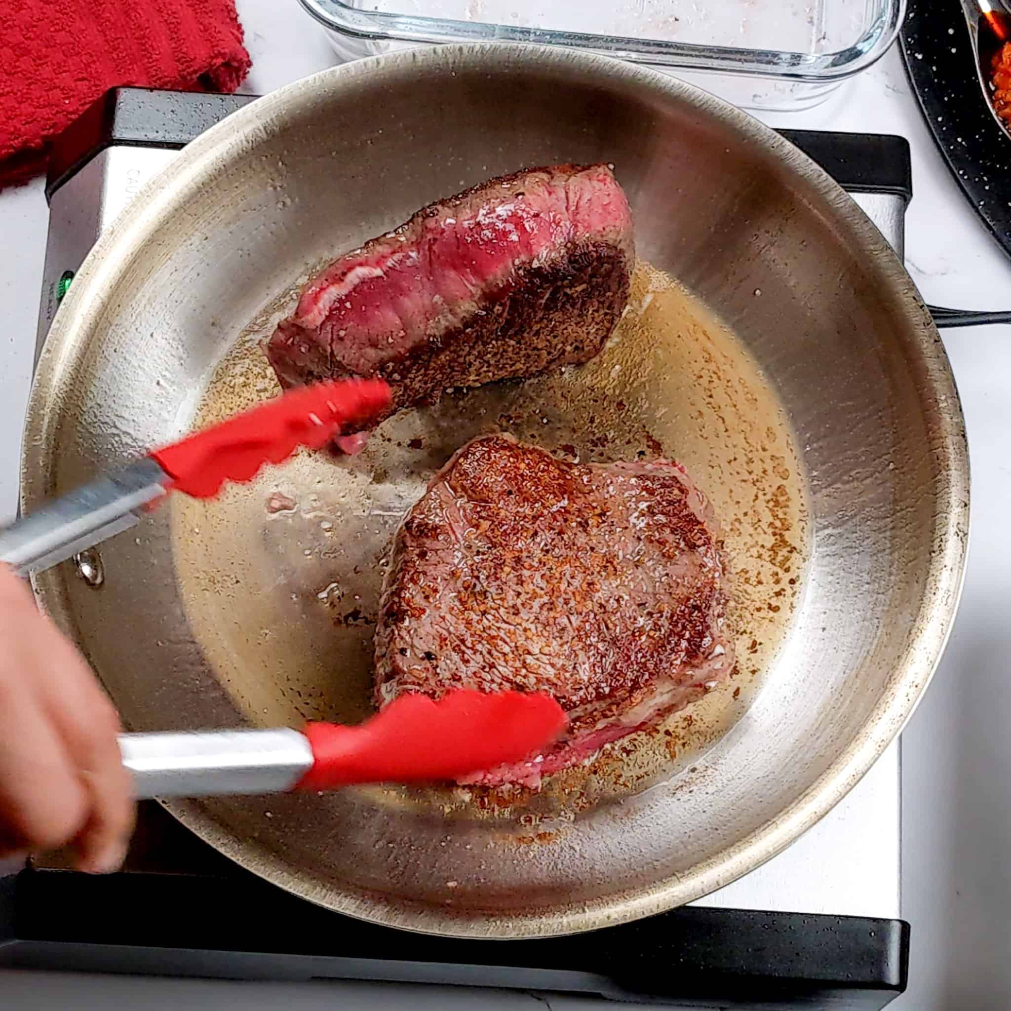 top sirloin steak being seared and flipped with a pair of kitchen-aid silicone tongs in an all-clad frying pan with oil for the Pan-Seared Sirloin Steak with Spicy Baharat Sauce recipe