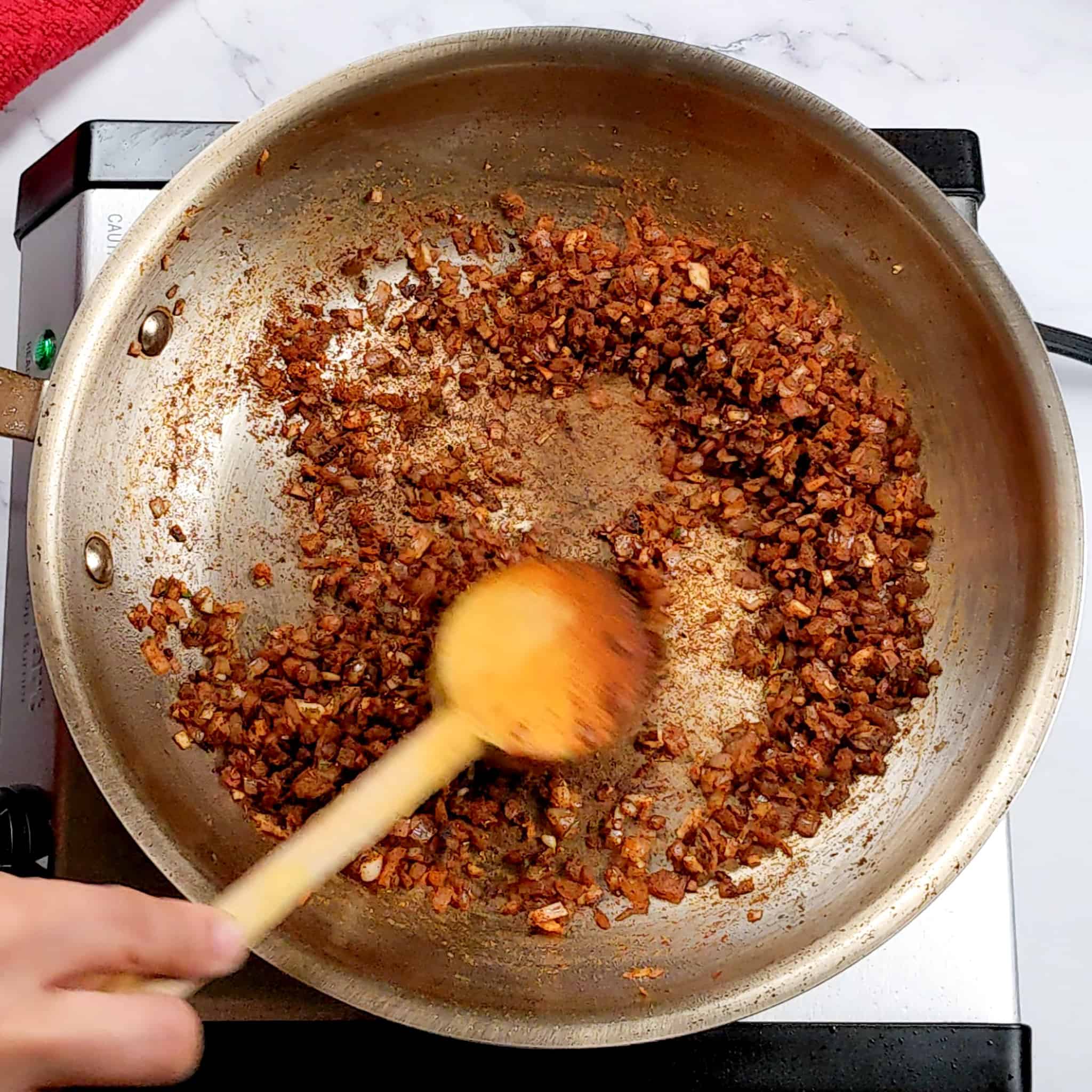 sauteed small diced shallots with chopped garlic and rosemary in a stainless steel frying pan mixed with Baharat seasoning being mixed with a wooden spoon for the sauteed small diced shallots with chopped garlic and rosemary in a frying pan recipe.