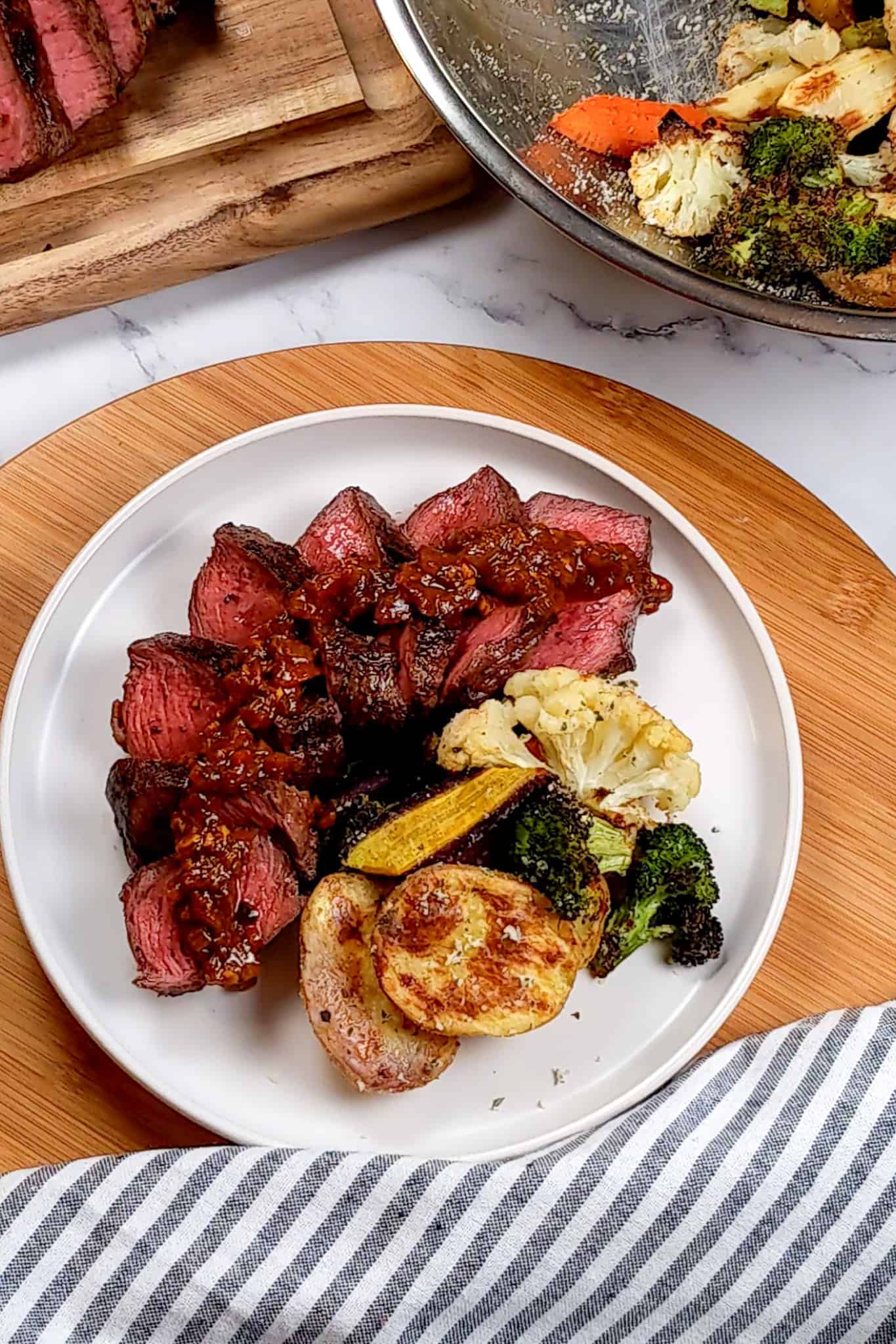 top view of the Pan-Seared Sirloin Steak with Spicy Baharat Sauce dish surrounded by a kitchen towel, bowl of roasted cheese tossed vegetables and sliced steak on a cutting board
