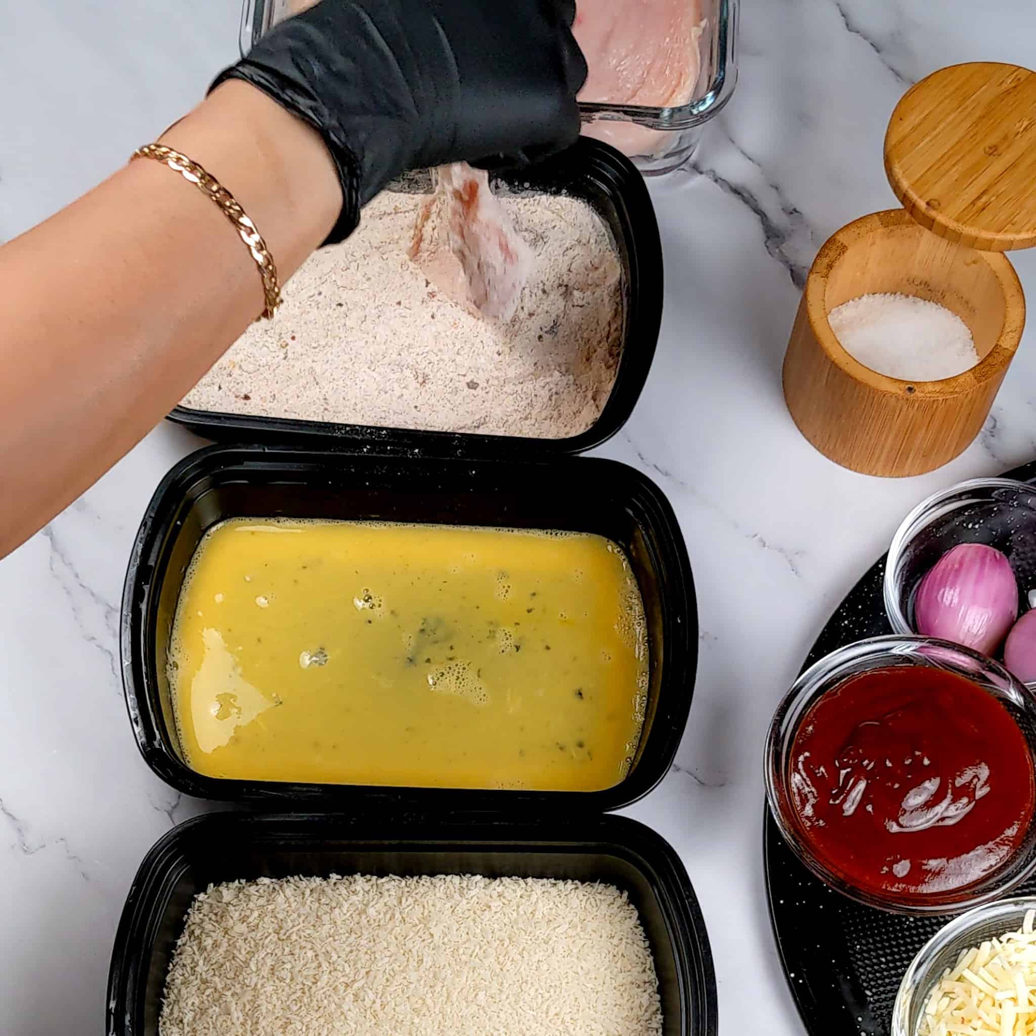 Chicken cutlet being dredged in seasoned flour in a container next to two other containers with beaten eggs and panko breadcrumbs.