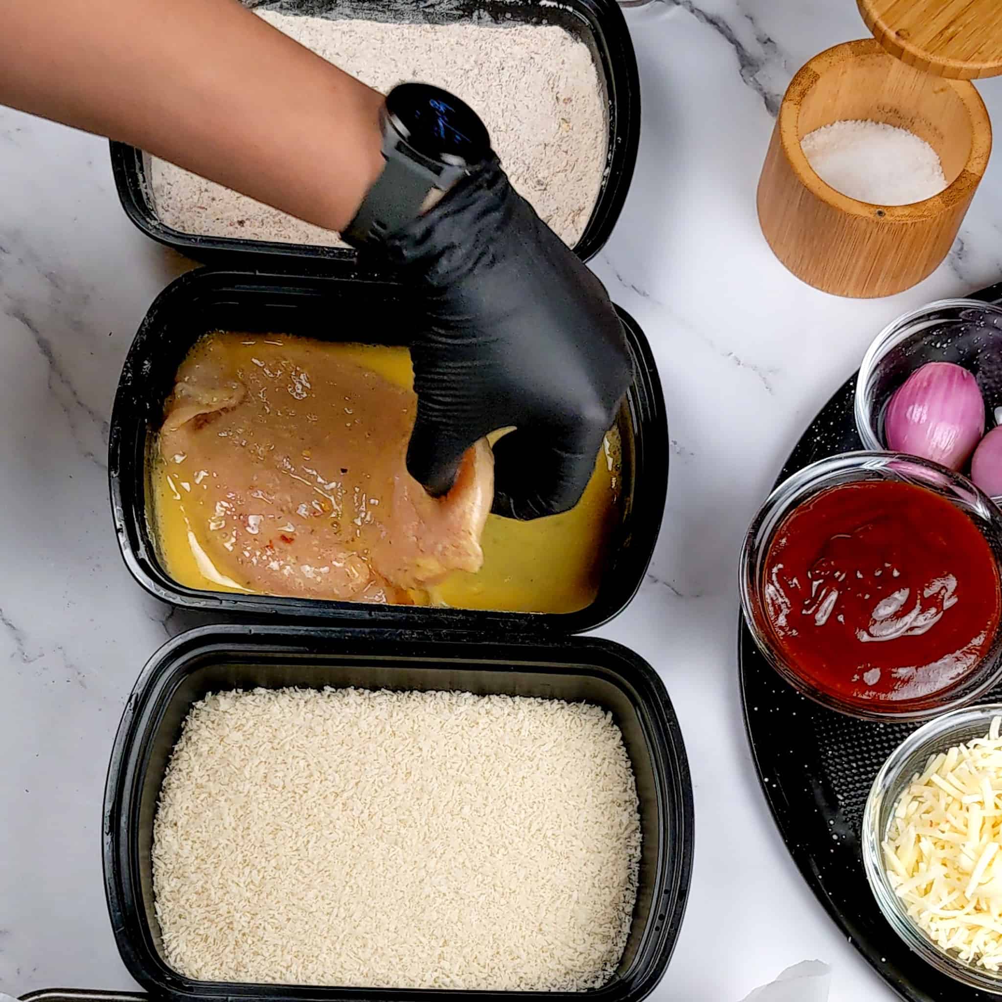 Chicken cutlet being dipped in beaten egg in container in between two other containers with seasoned flour and panko breadcrumbs.