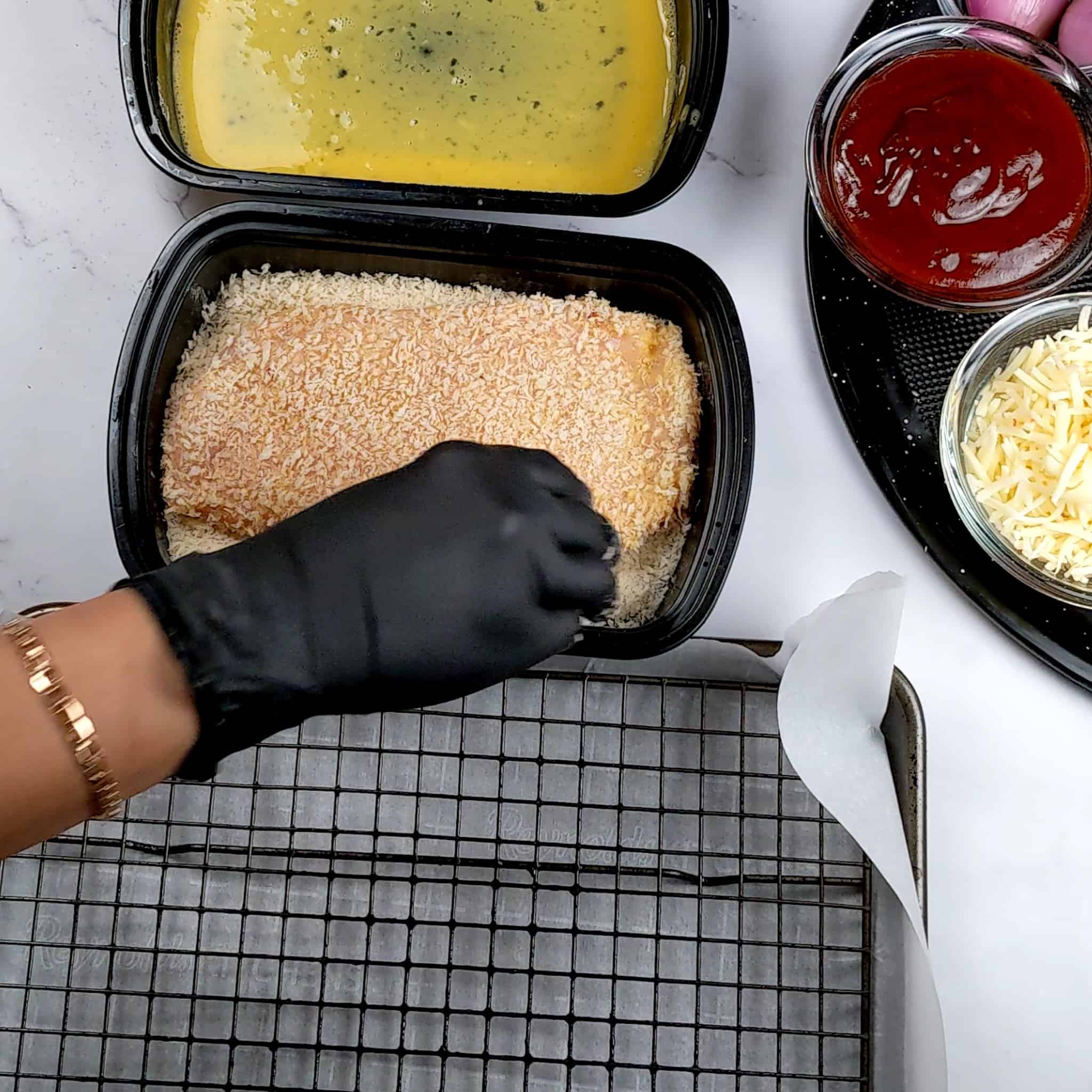 Chicken cutlet being coated in panko breadcrumbs in a container next two other containers with seasoned flour and beaten eggs.