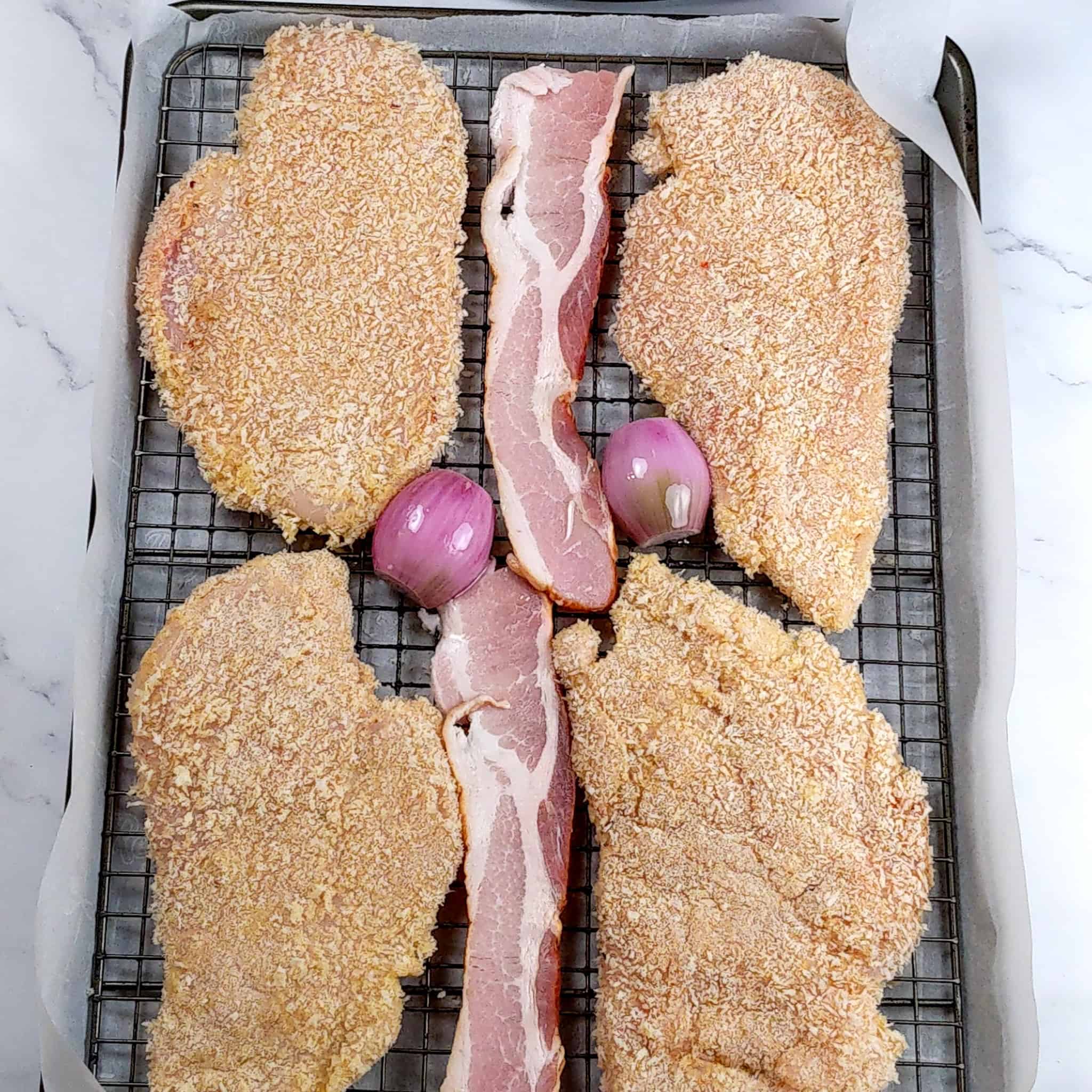 breaded chicken cutlets on a sheet pan lined with parchment paper on a rack, with bacon strips and halved shallots in between for the Easy Baked Cheesy Crispy Barbeque Chicken Cutlets.