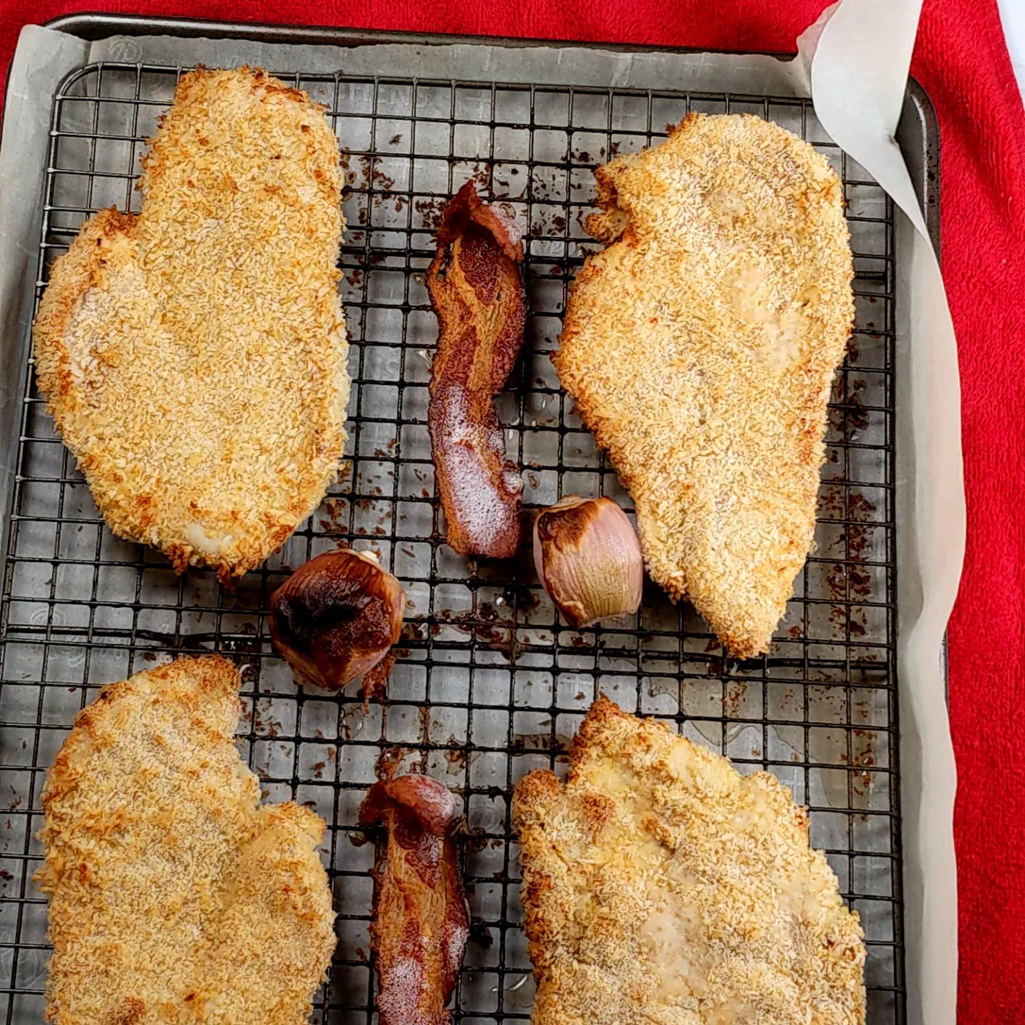 Cooked breaded chicken cutlets, bacon and shallots on a rack on a sheet pan lined with parchment paper.