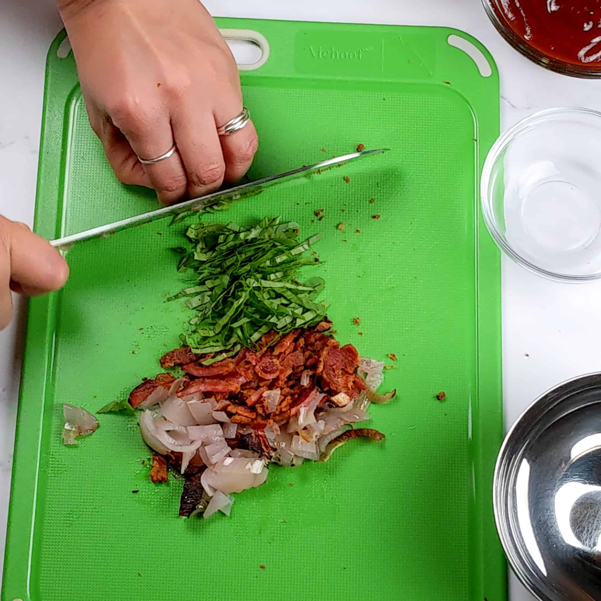 Basil being chopped next to chopped bacon and roasted shallots on a cutting board for the Easy Baked Cheesy Crispy Barbeque Chicken Cutlets recipe.
