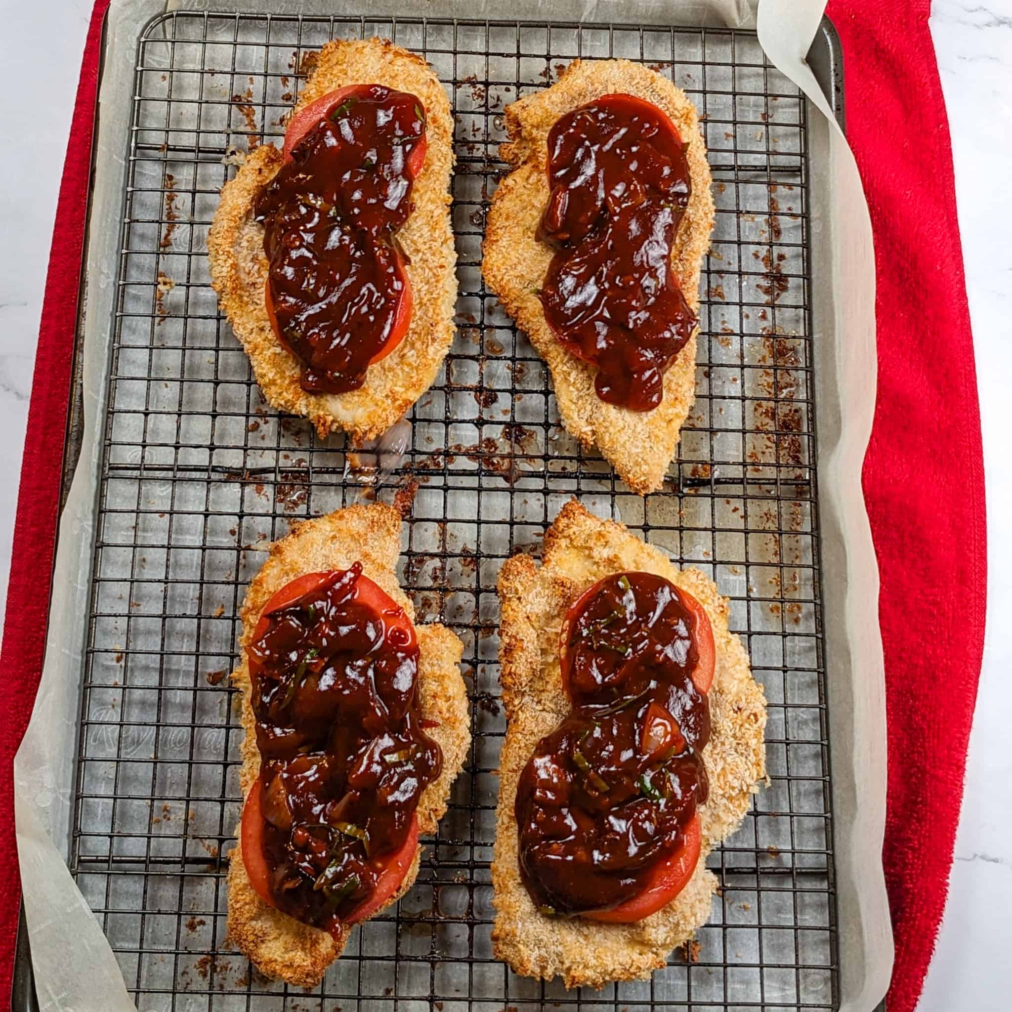 Baked chicken cutlets topped with sliced tomatoes and barbeque sauce on a sheet pan with a rack.