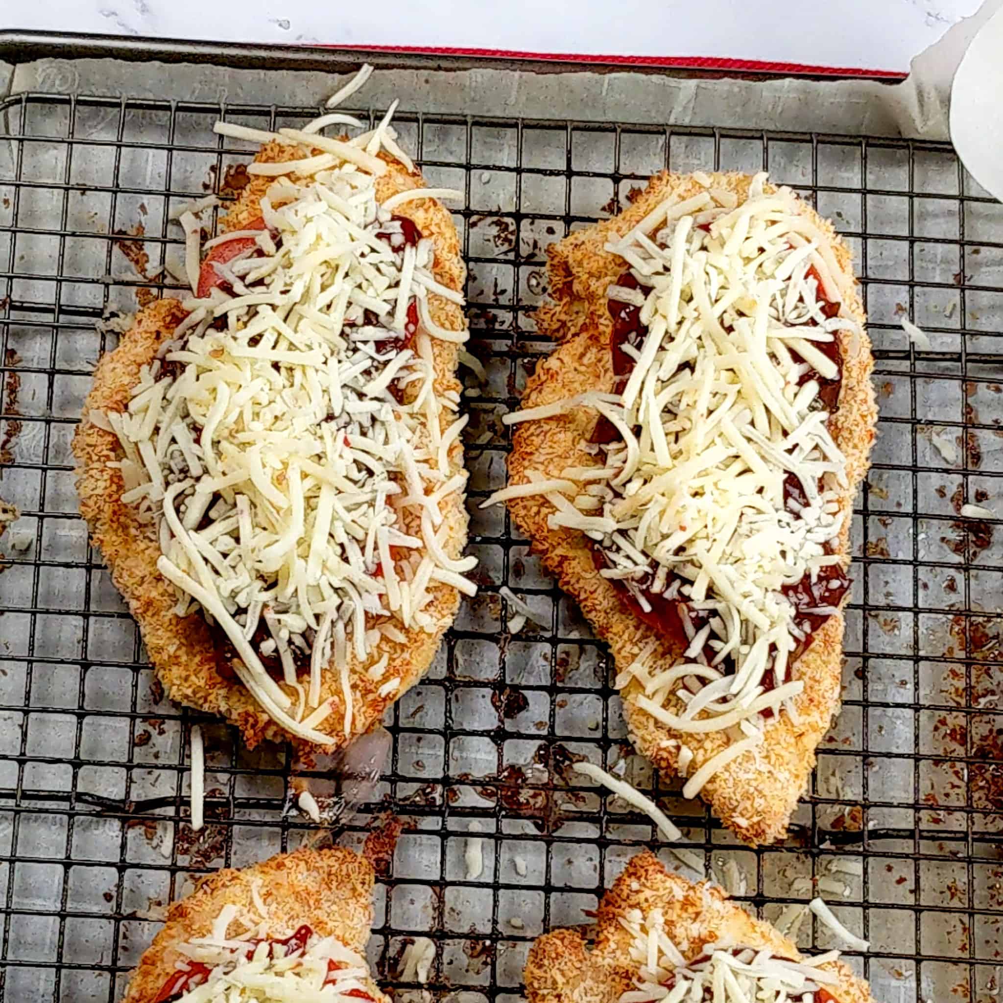 Baked chicken cutlets topped with sliced tomatoes, barbeque sauce, and shredded pepper jack cheese on a sheet pan with a rack.