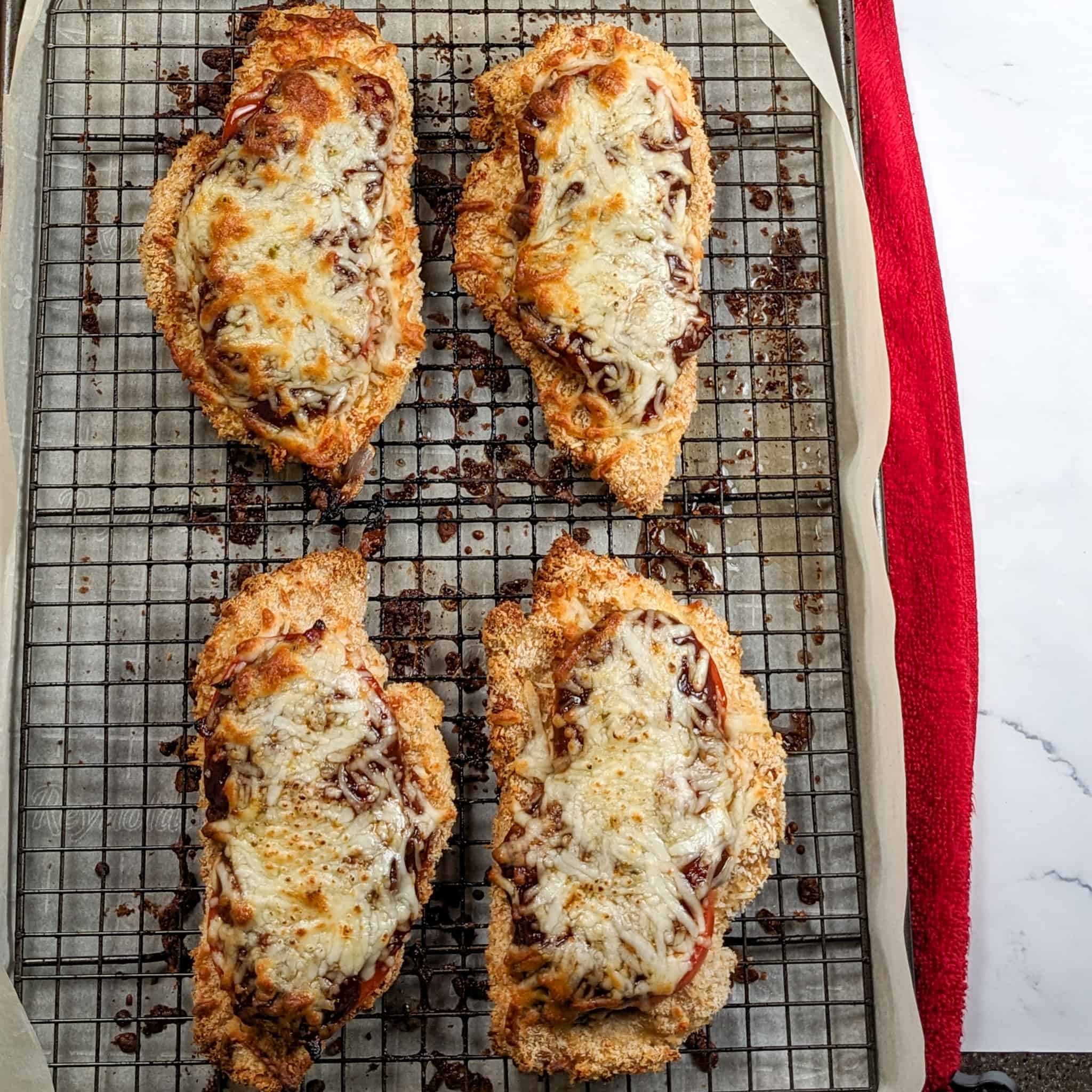 Breaded chicken cutlets topped with sliced tomatoes, barbeque sauce, and shredded pepper jack cheese baked on a sheet pan with a rack.