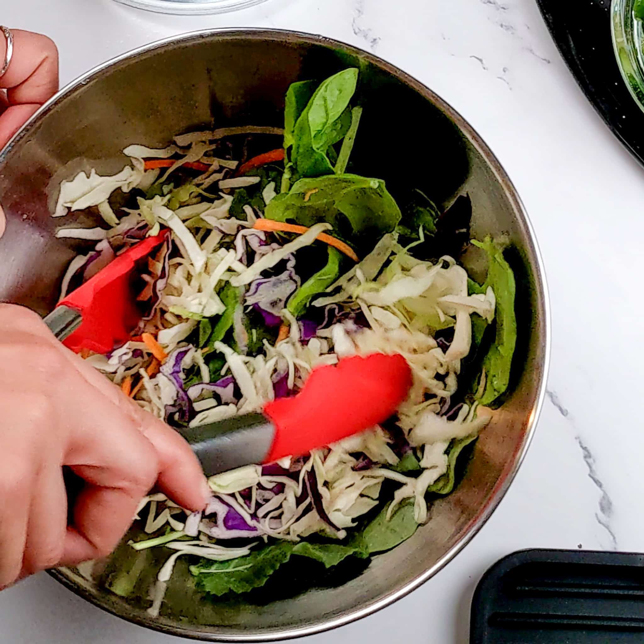 veggie slaw with mixed baby greens being mixed together with a pair of silicone tongs in a stainless steel mixing bowl
