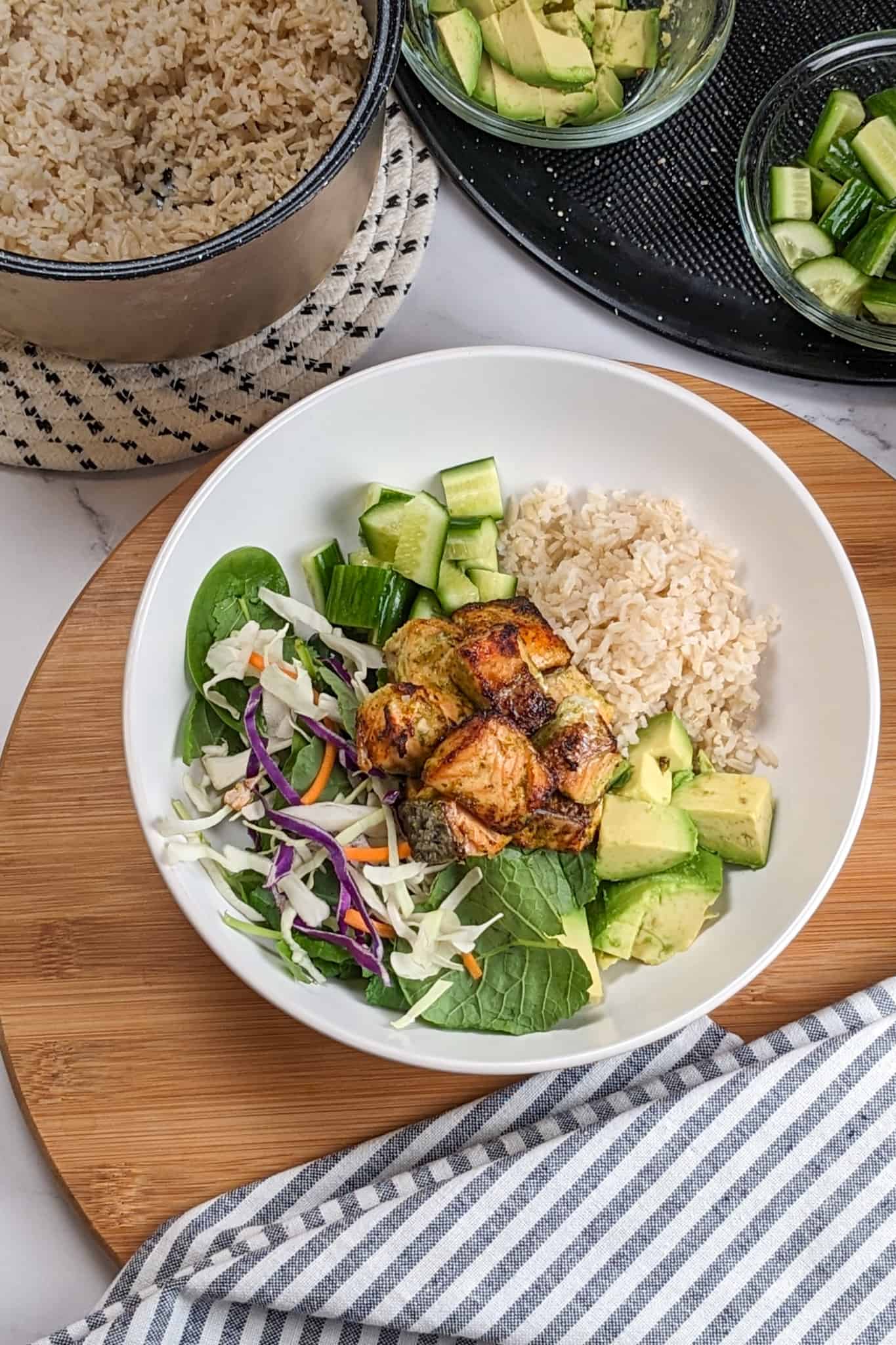 top view of the Air Fryer Spicy Cilantro Lime Salmon Salad Rice Bowl dish on a wooden lazy Susan next to a kitchen towel, rice cooker container and remaining rice bowl ingredients in containers on a large pizza pan