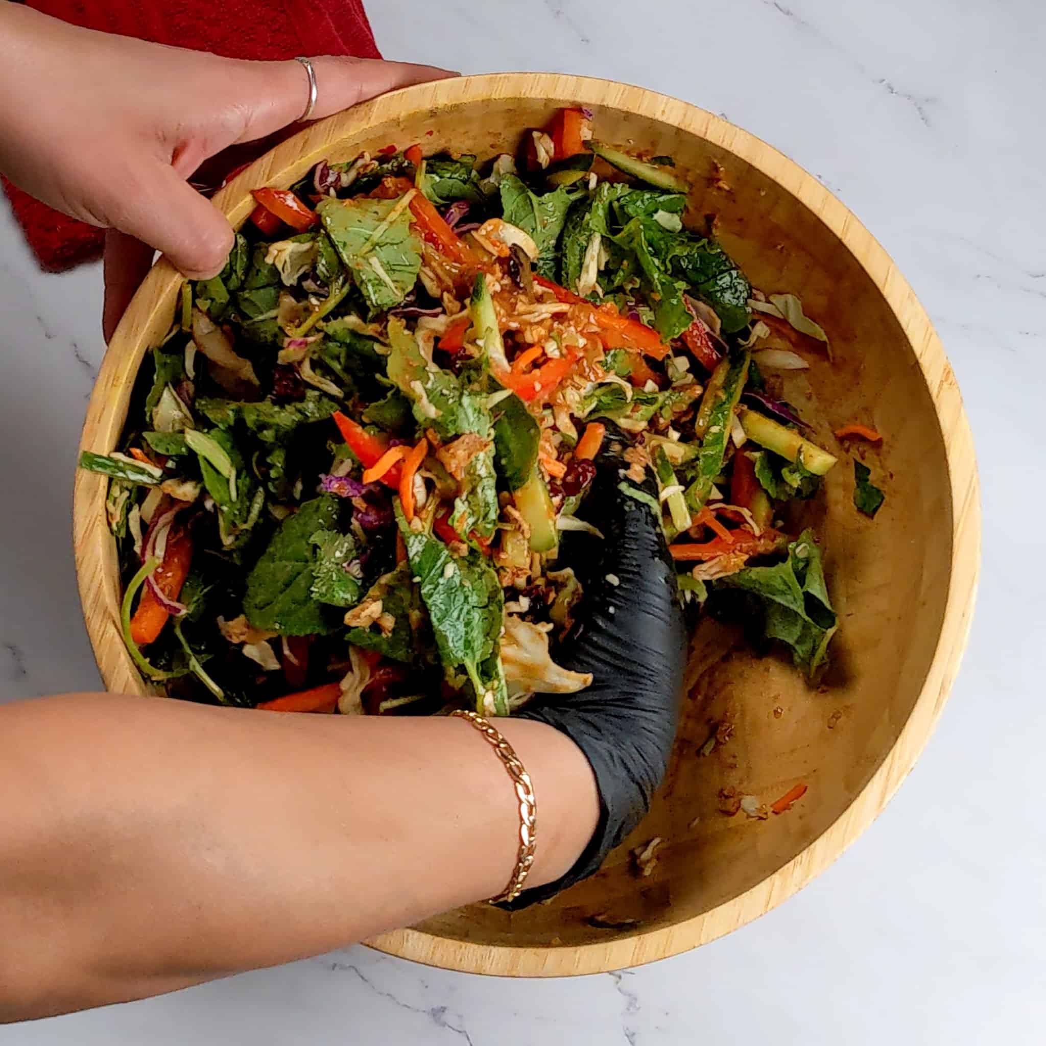 a gloved hand gently mixing the vegetables and greens in the wooden bowl.