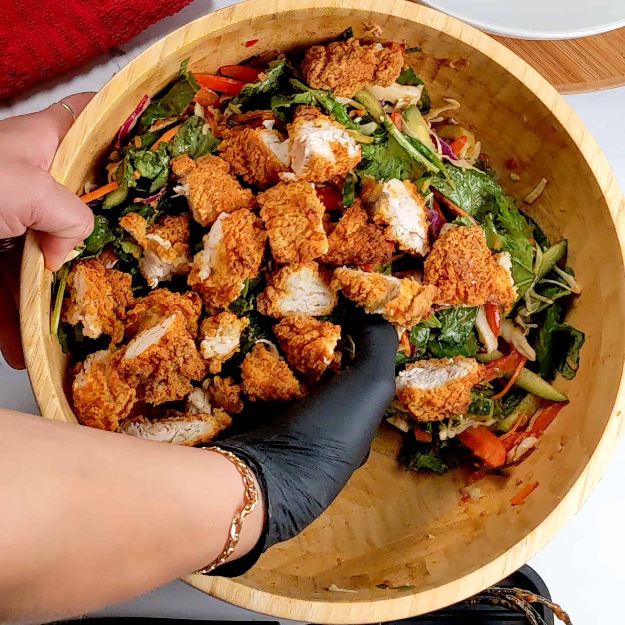 chopped chicken tenders being gently tossed into the salad by a gloved hand in a wooden bowl for the Sweet Chili Peanut Chicken Tenders Salad