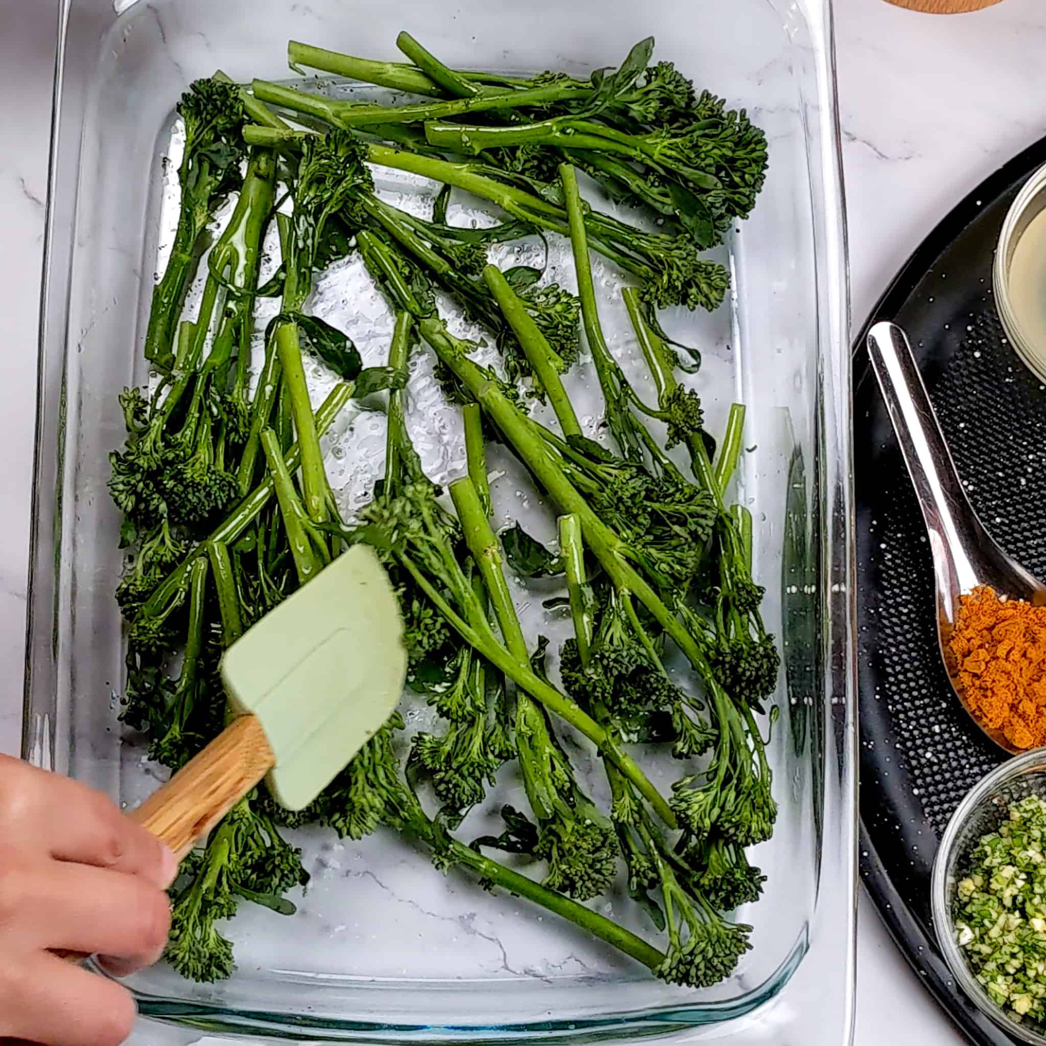 broccolini being tossed with a silicone wooden handle spatula in a glass rectangle baking dish