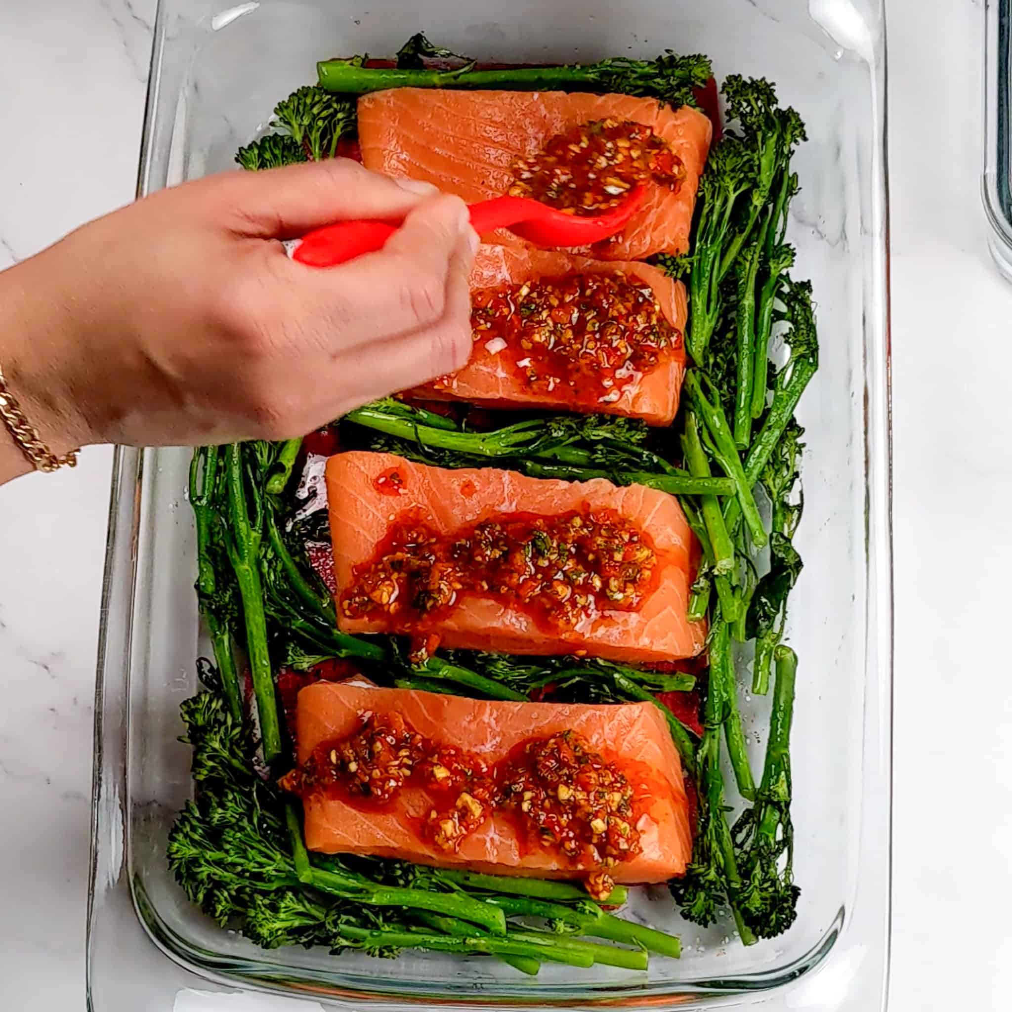 Calabrian pepper basil sauce being spooned onto the salmon fillets with a silicone spoon in a rectangle large glass baking dish filled with broccolini for the Calabrian Pepper Basil Baked Salmon and Broccolini recipe.
