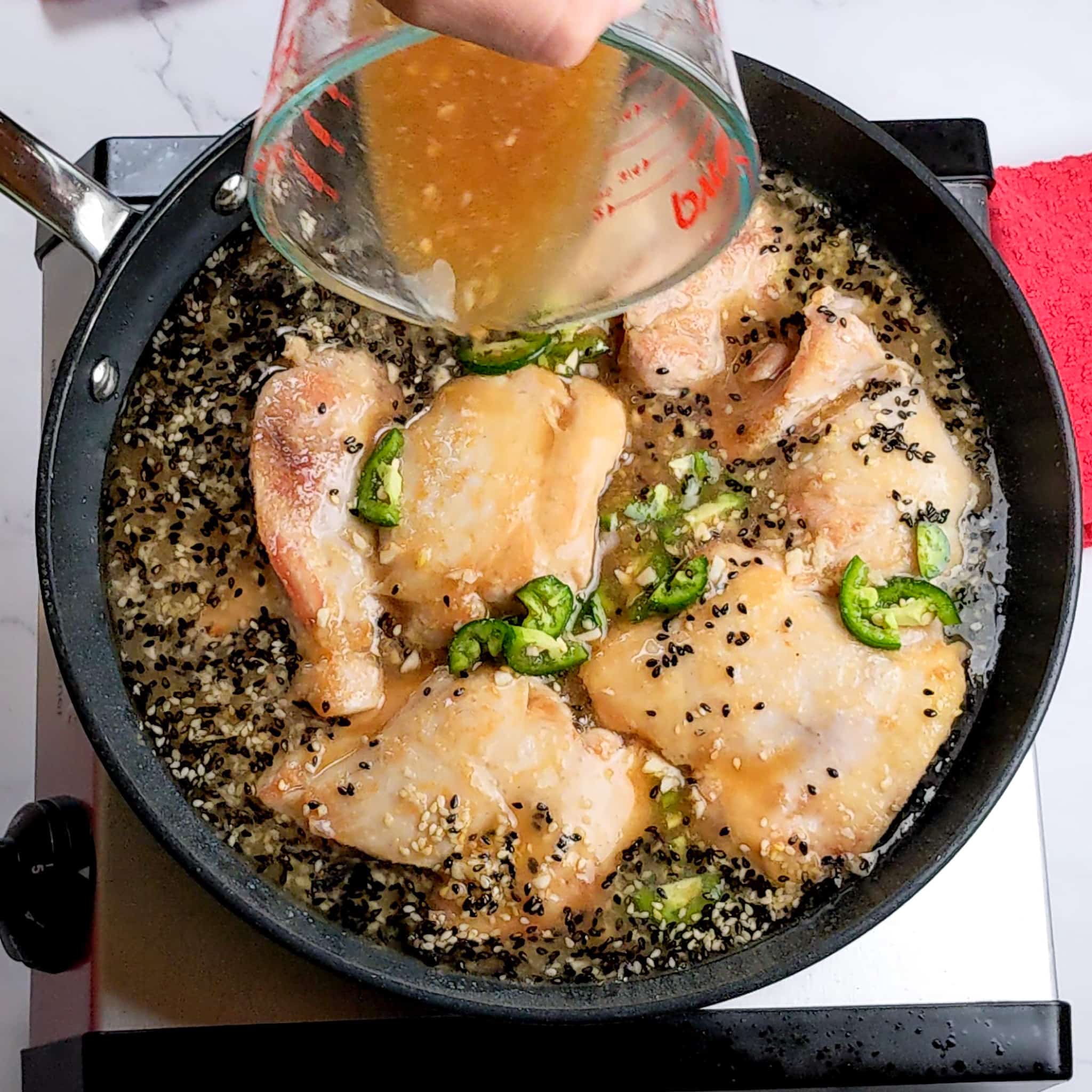 seasoned broth being poured from a measuring cup into the toasted sesame seed and chicken mixture in a non stick frying pan