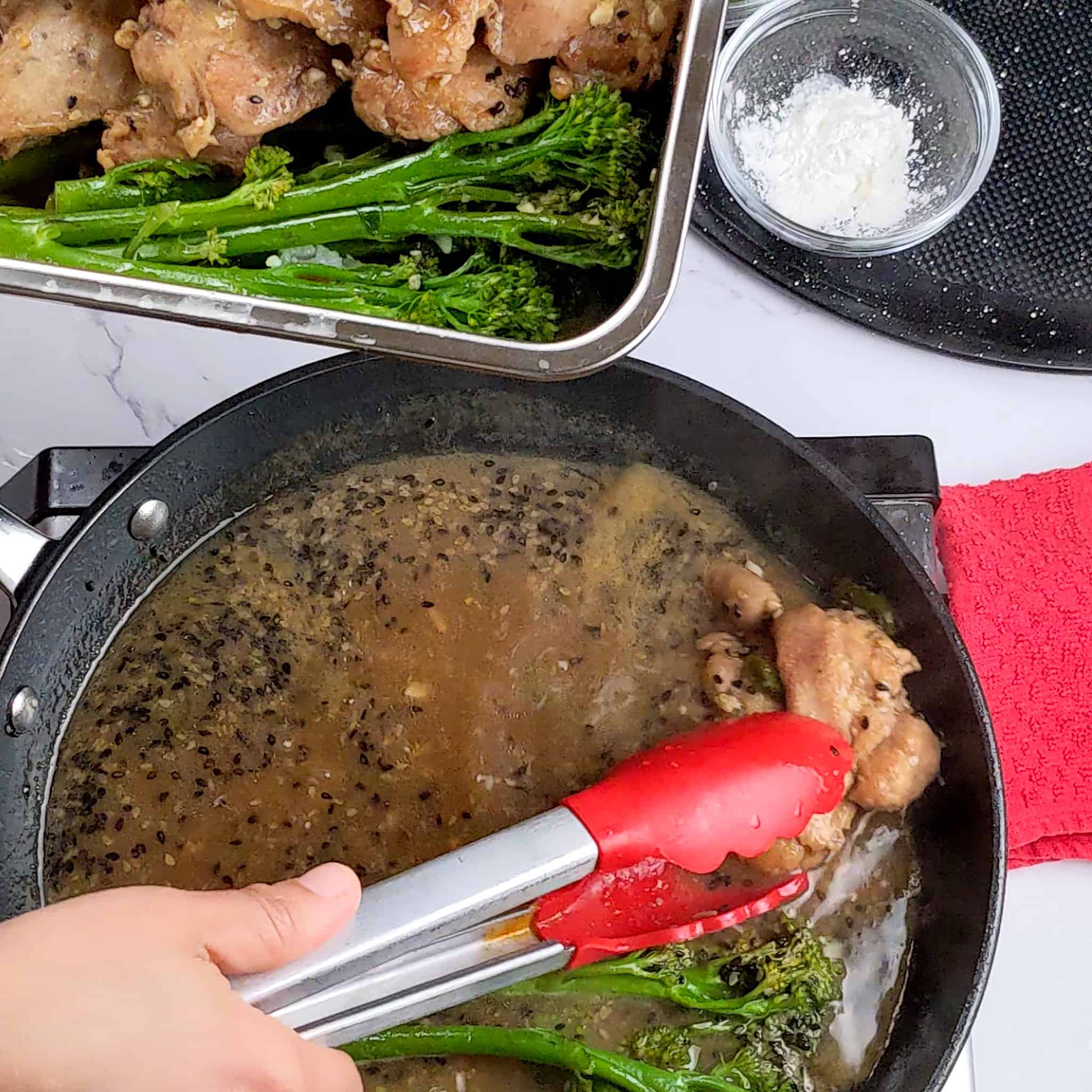 the cooked chicken and broccolini being removed from a non-stick frying pan with a pair of silicone tipped stainless steel tongs