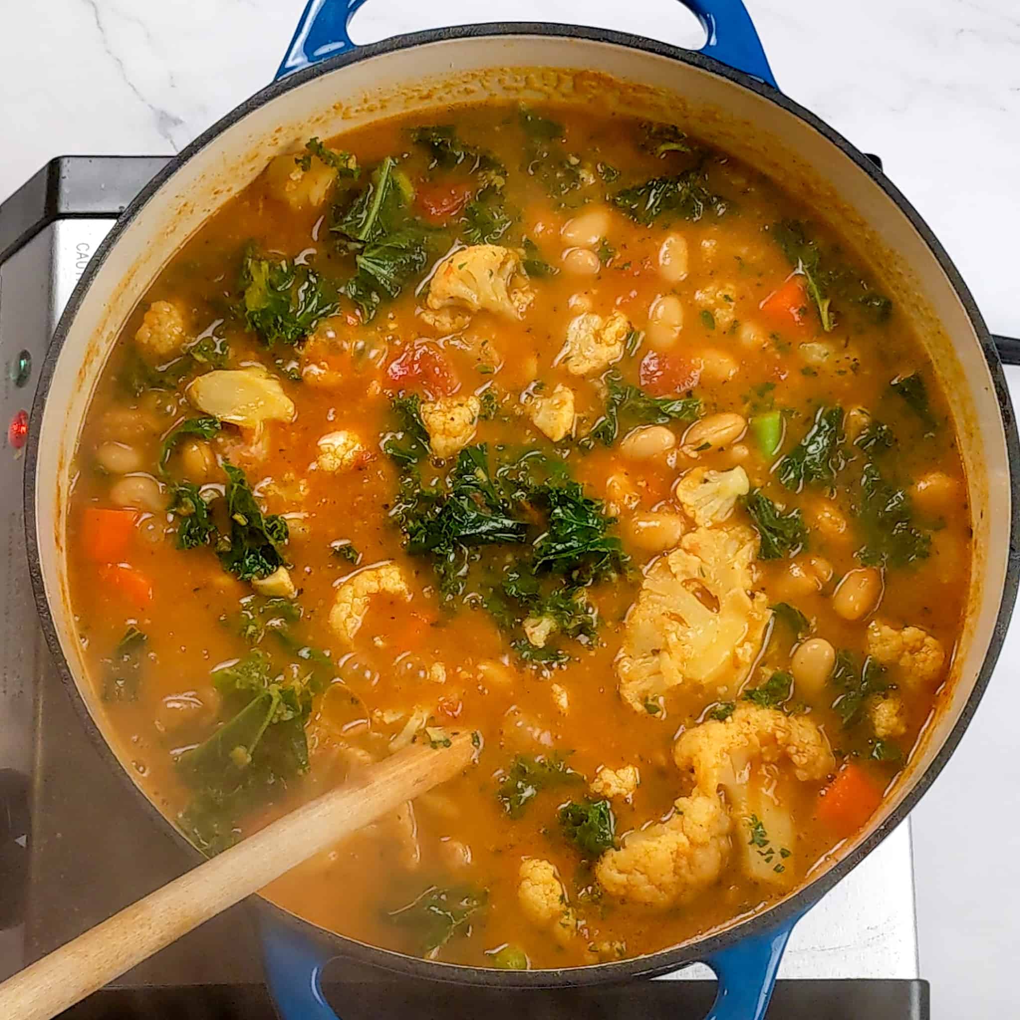 kale, cannellini beans, cauliflower and carrots floating a tomato based broth in a dutch oven with a wooden spoon