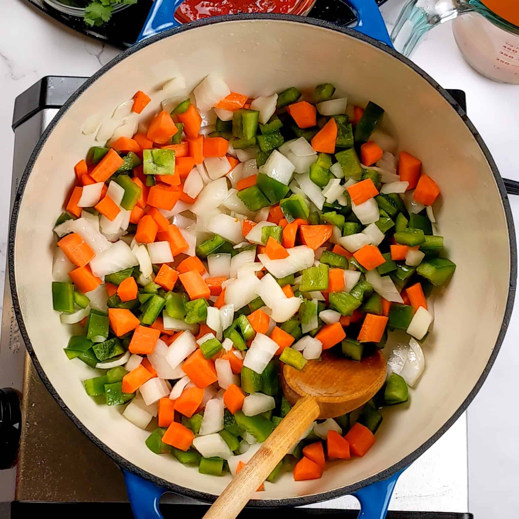 a dutch oven with diced onion, green bell peppers and carrots combined with a wooden spoon