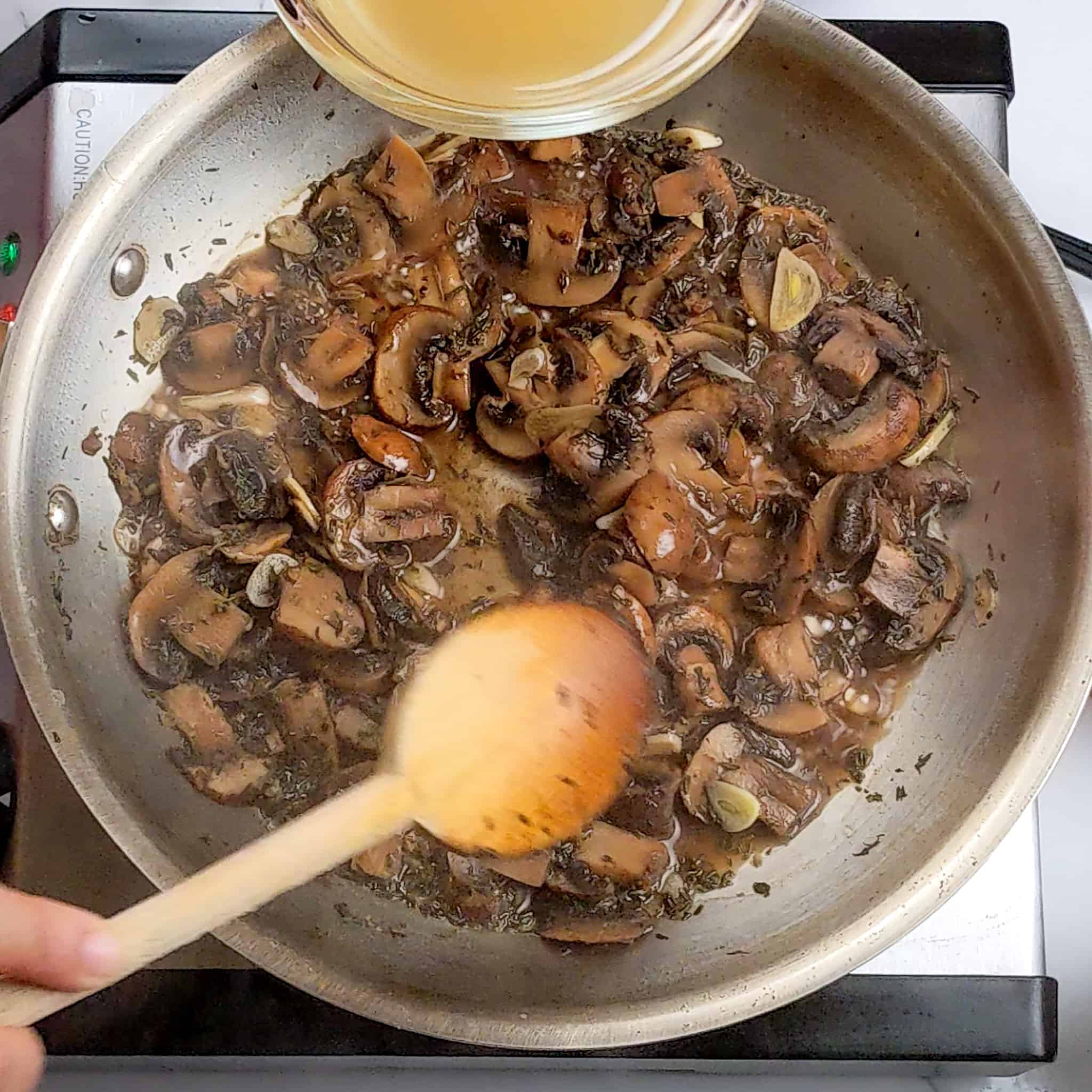 rum soaked sauteed sliced mushrooms cooking in chicken broth and being mixed with a wooden spoon