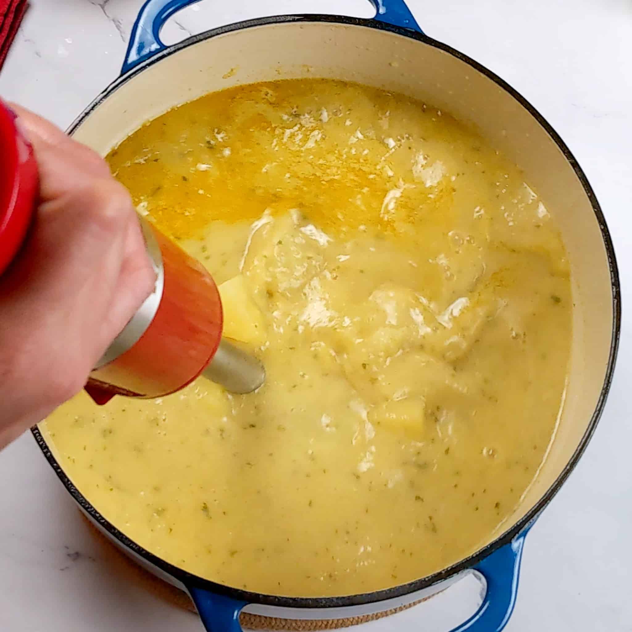 potato chunks in a seasoned broth being blended with a kitchenaid immersion handheld wireless blender in a loge dutch oven for the Best Fragrant Creamy Potato Coconut Curry Fish Soup recipe