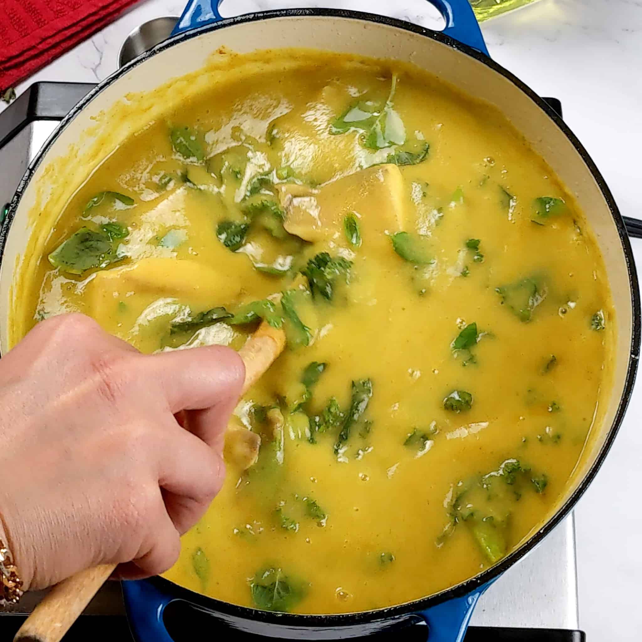 fresh tilapia fish chunks added to the creamy curry potato coconut and kale soup for the Best Fragrant Creamy Potato Coconut Curry Fish Soup recipe being gently stirred with a wooden spoon