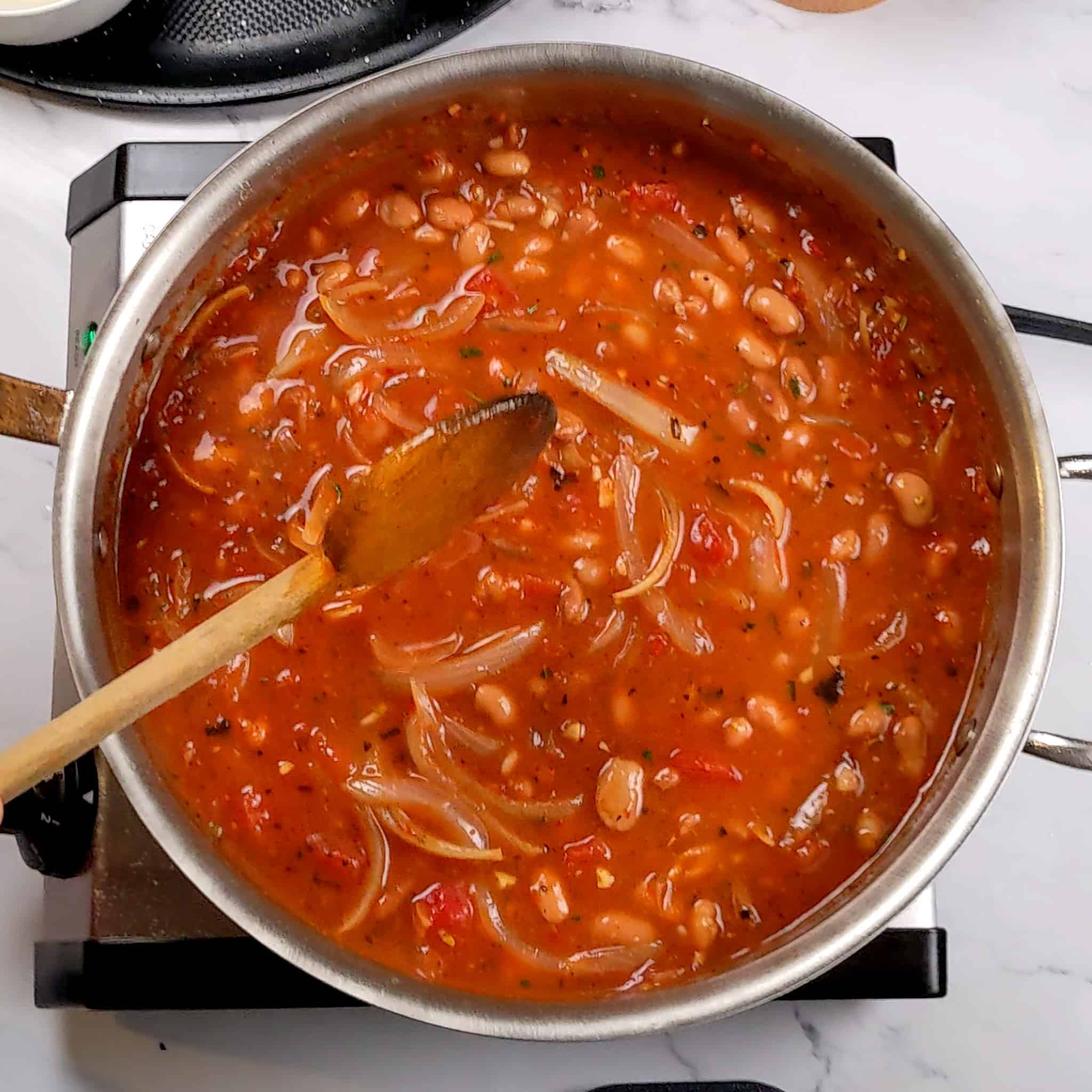 tomato-based onion and bean saute being mixed with a wooden spoon in an all-clad stainless steel saute pan