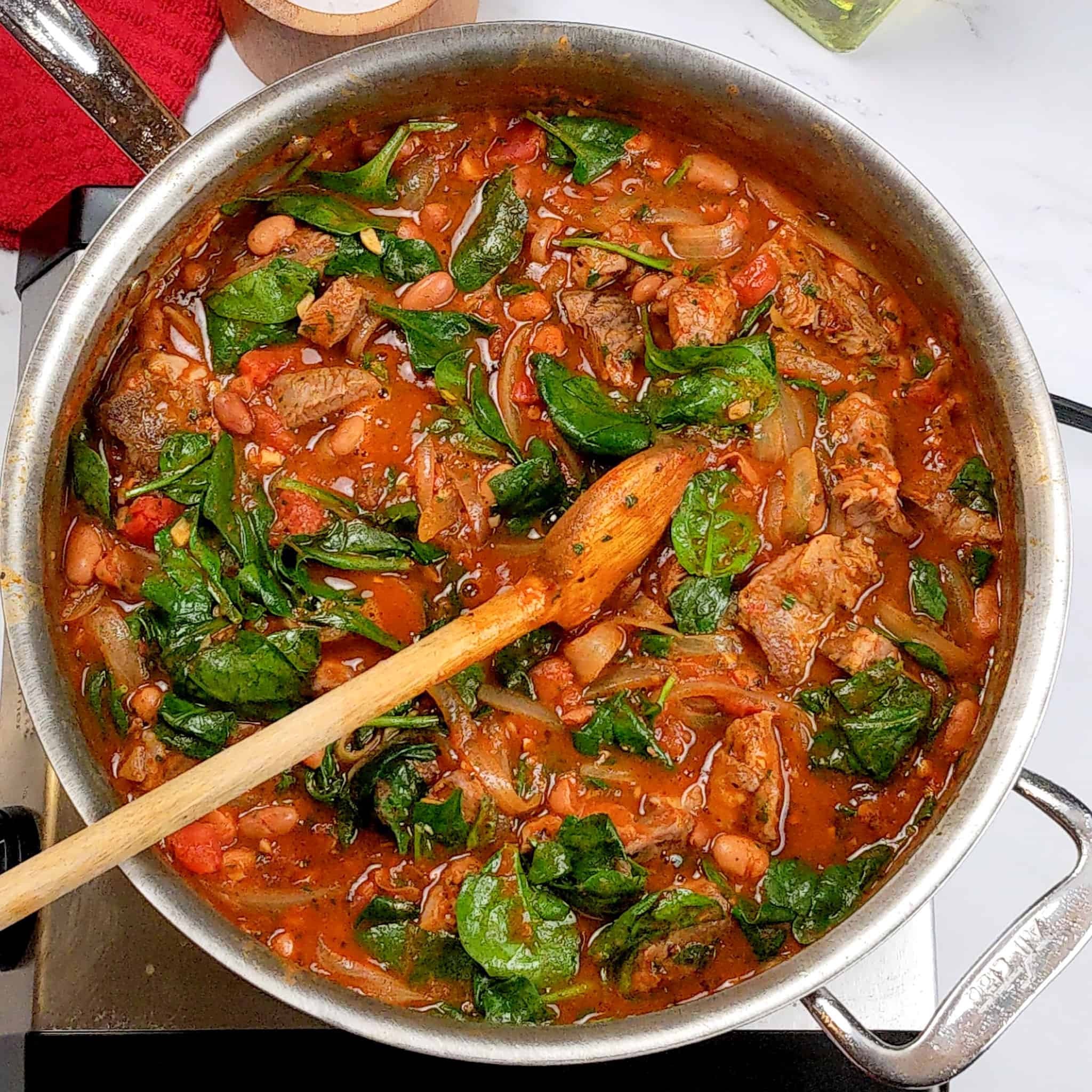 the Quick and Easy Lean Baharat Spiced Bean and Lamb Stew being mixed with a wooden spoon in an all-clad stainless steel saute pan