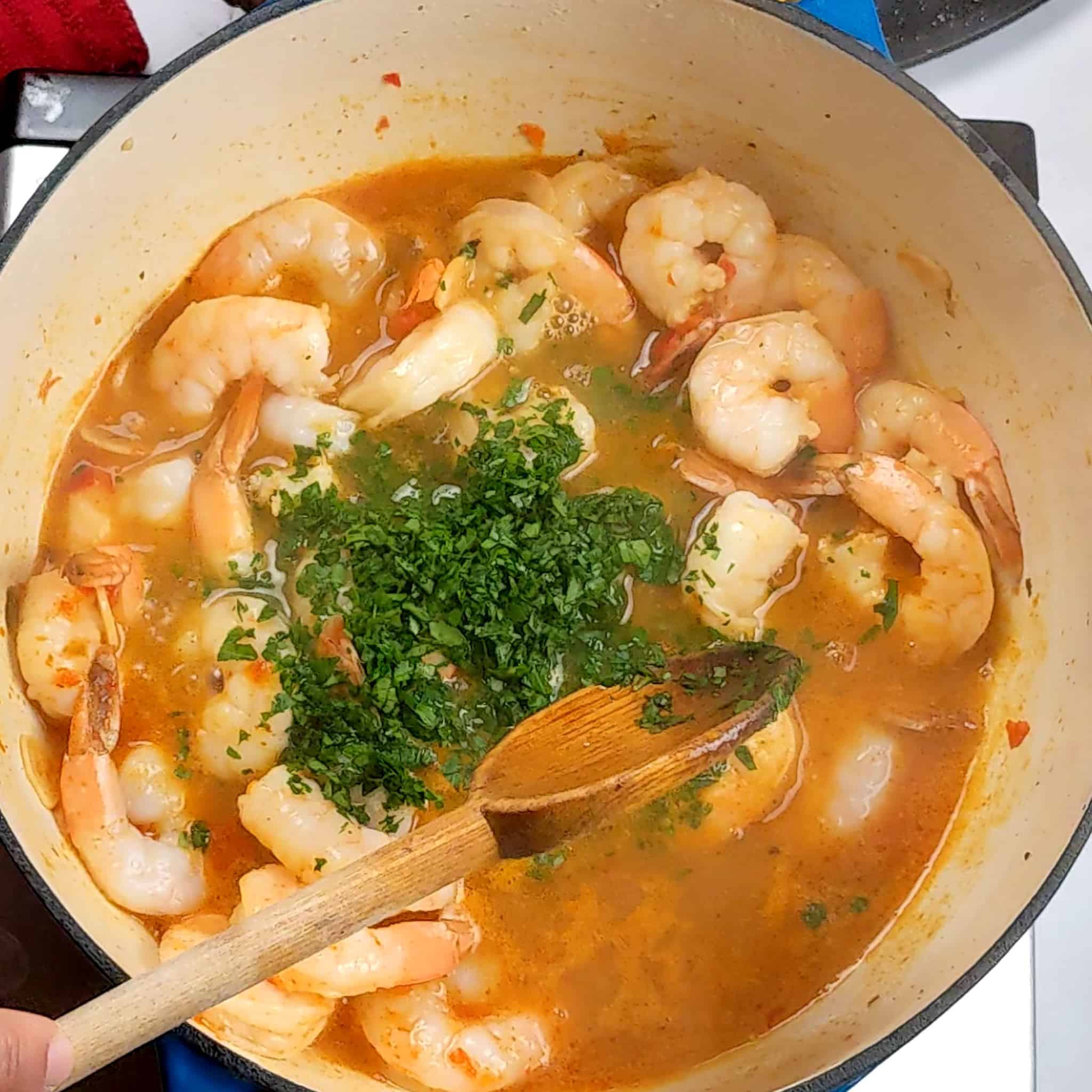 cooked shrimp in garlic chili broth with chopped parsley drop into the middle about to be mixed with a wooden spoon