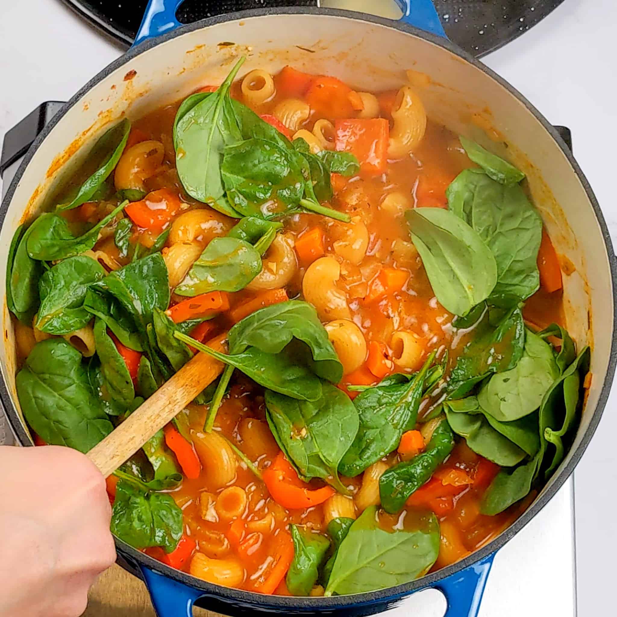 spinach added and s stirred with a wooden spoon to a pot of macaroni pasta and red bell peppers in a rich tomato broth in a dutch pan
