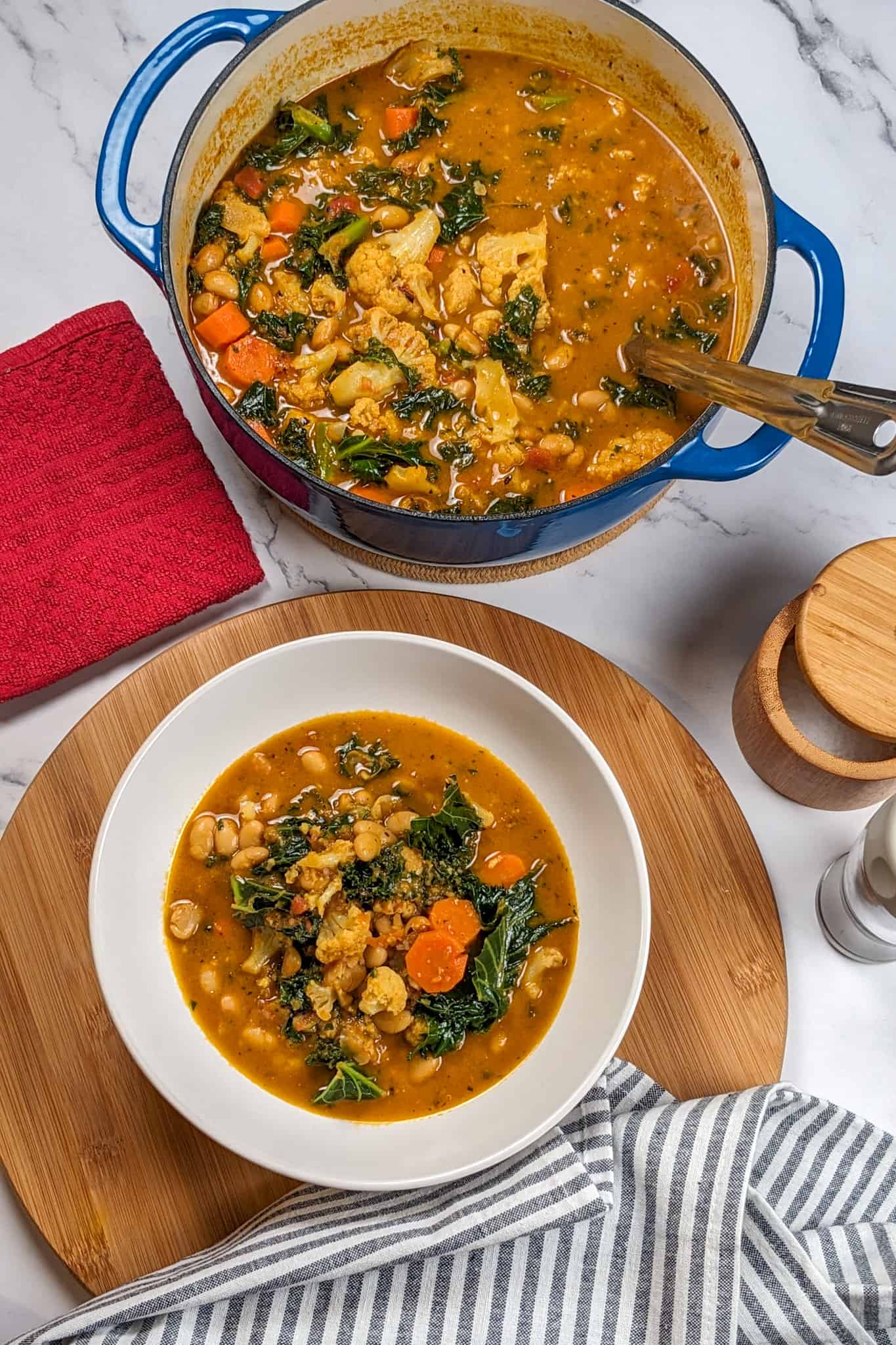 top view of the bean kale tomato stew in a wide rim bowl on a wooden lazy susan with a kitchen towel resting next to it with the dutch oven with the remaining stew with a ladle