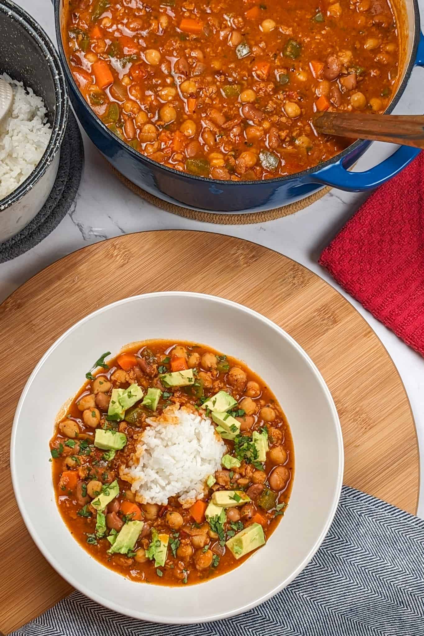 top view of the Vegetarian Smoky Chipotle Chorizo and Bean Stew surrounding cooked rice in a wide rim bowl on a wooden lazy susan next to a kitchen towel and a dutch pot of the remaining chili with a stainless steel ladle and a container of steamed jasmine rice